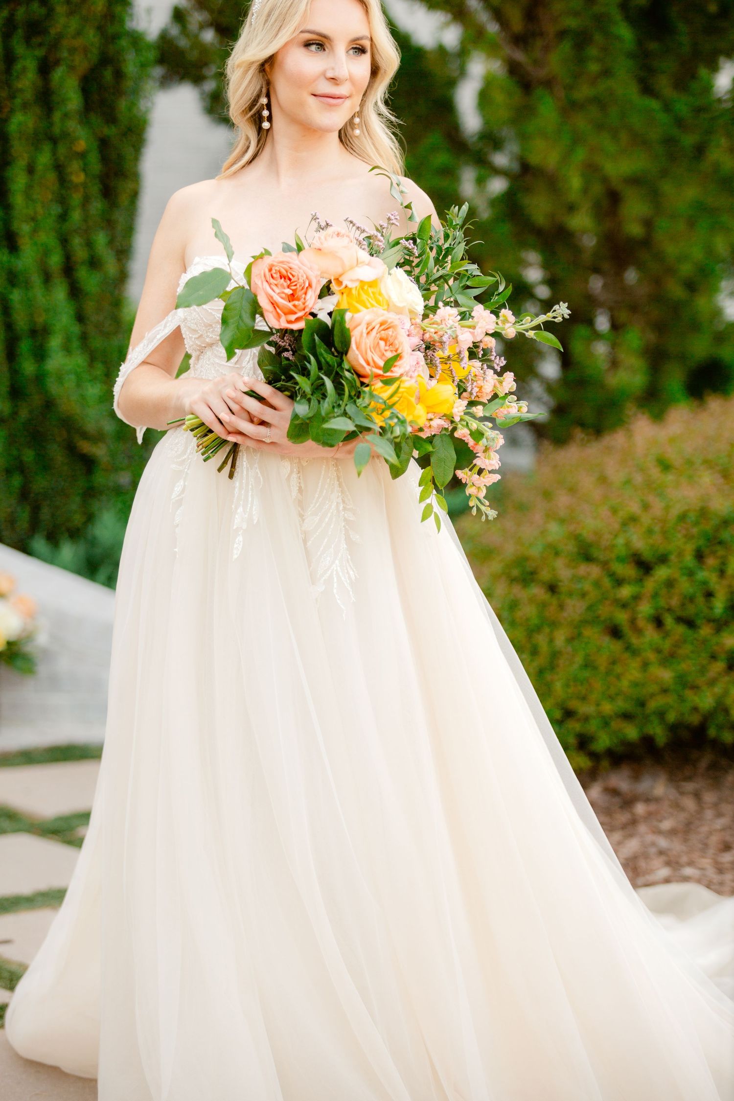 A bride in a flowing white gown holds a vibrant coral and yellow floral bouquet against a lush garden backdrop.
