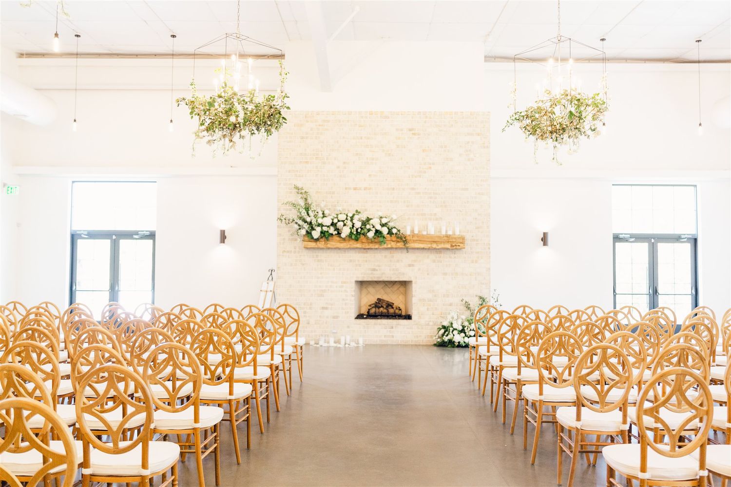 Modern white wedding ceremony space with wooden chairs arranged in rows and floral arrangements hanging from the walls.