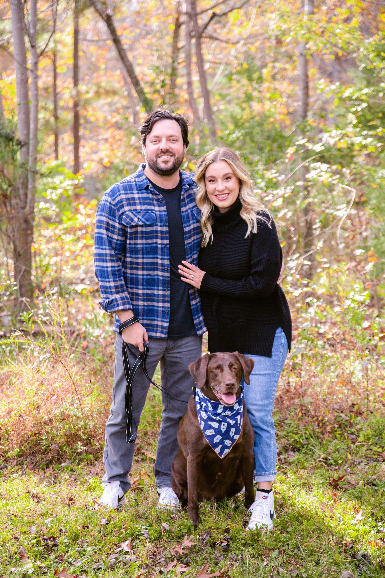 Couple with their chocolate lab dog posing in autumn woods with colorful foliage.