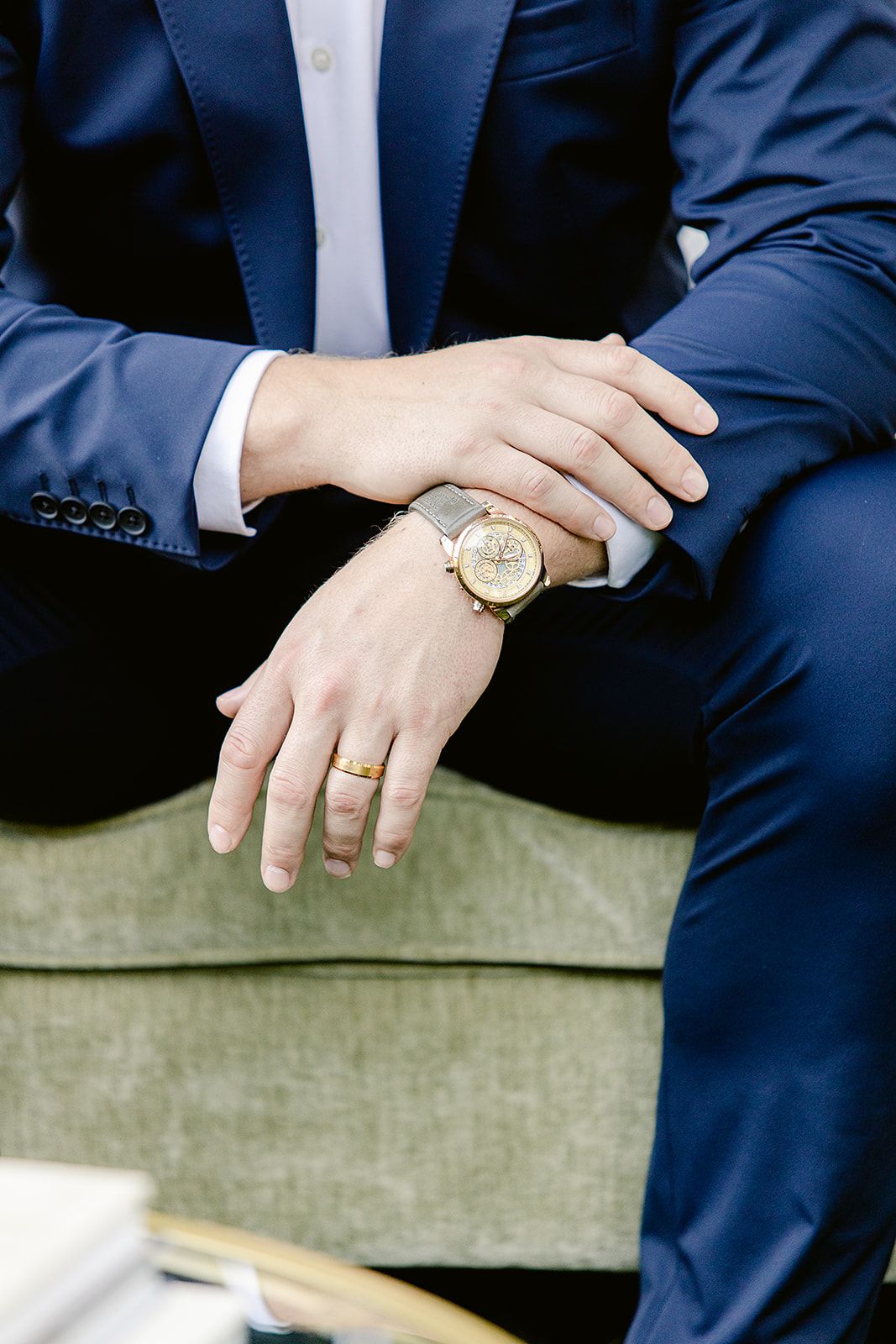 Detail shot of hands with wedding rings and gold watch against a navy suit and green sofa background.