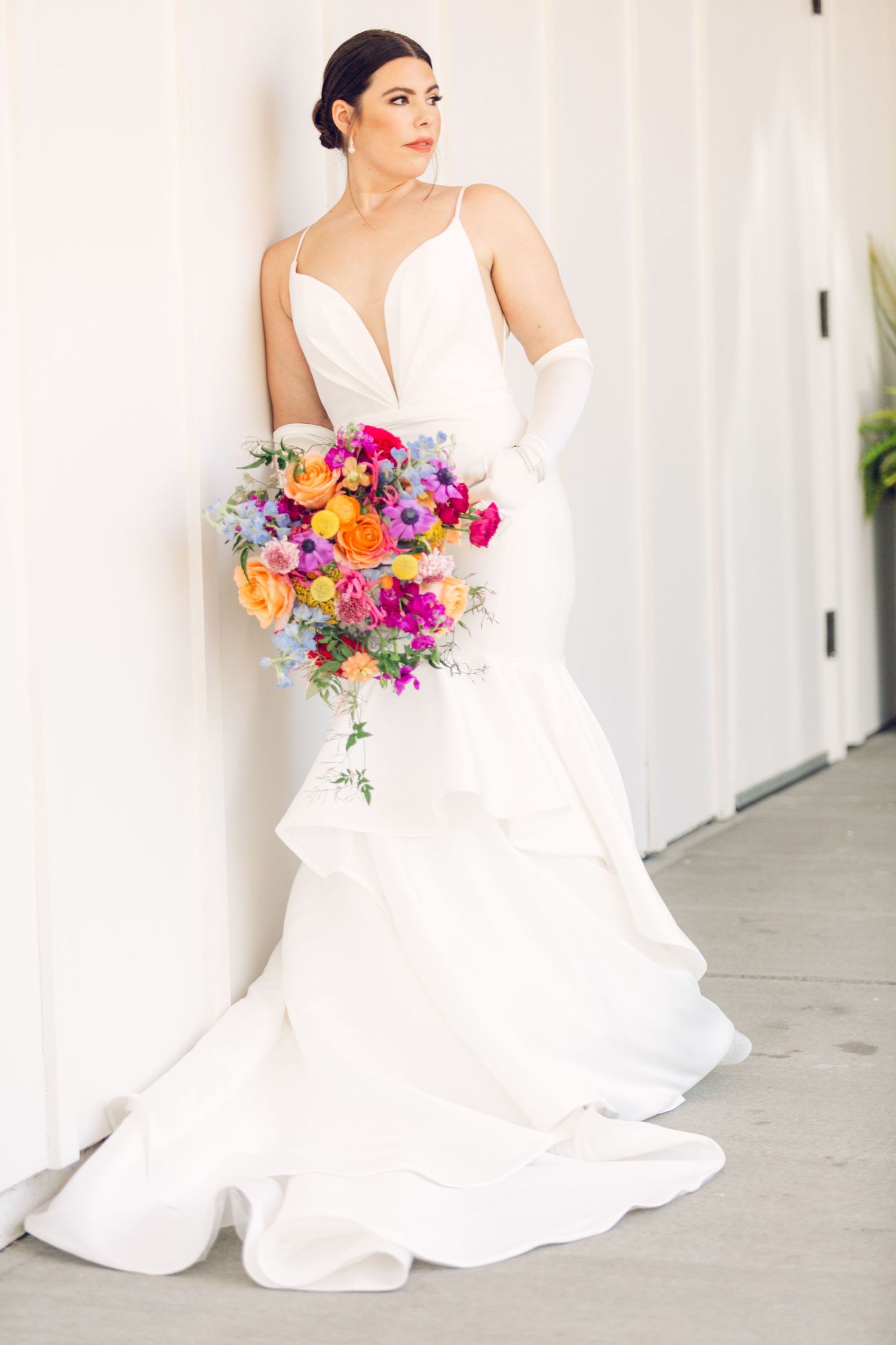 Modern bride in flowing white dress holding a bright multicolored bouquet against a white backdrop.