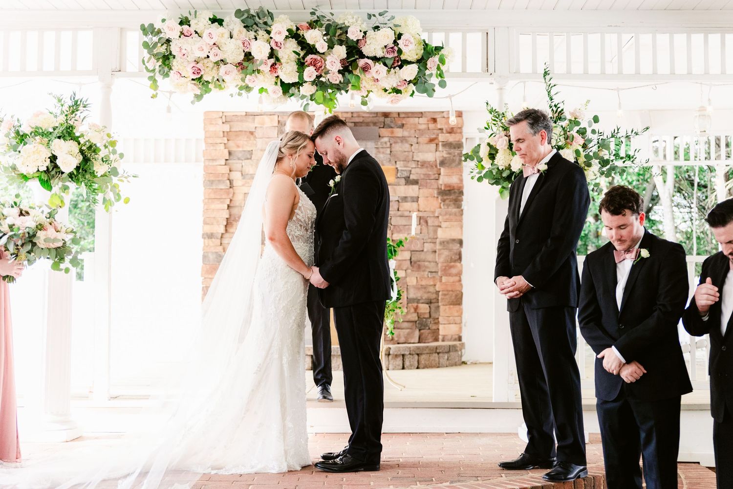 Wedding ceremony in progress under a floral arch with brick backdrop and white covered porch.
