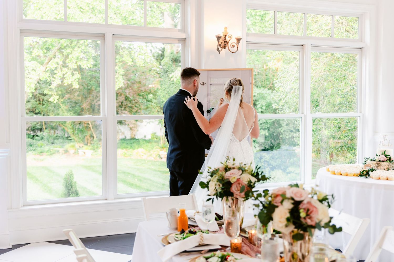 Bright wedding reception hall with white decor, tall windows, and floral arrangements on dining tables.