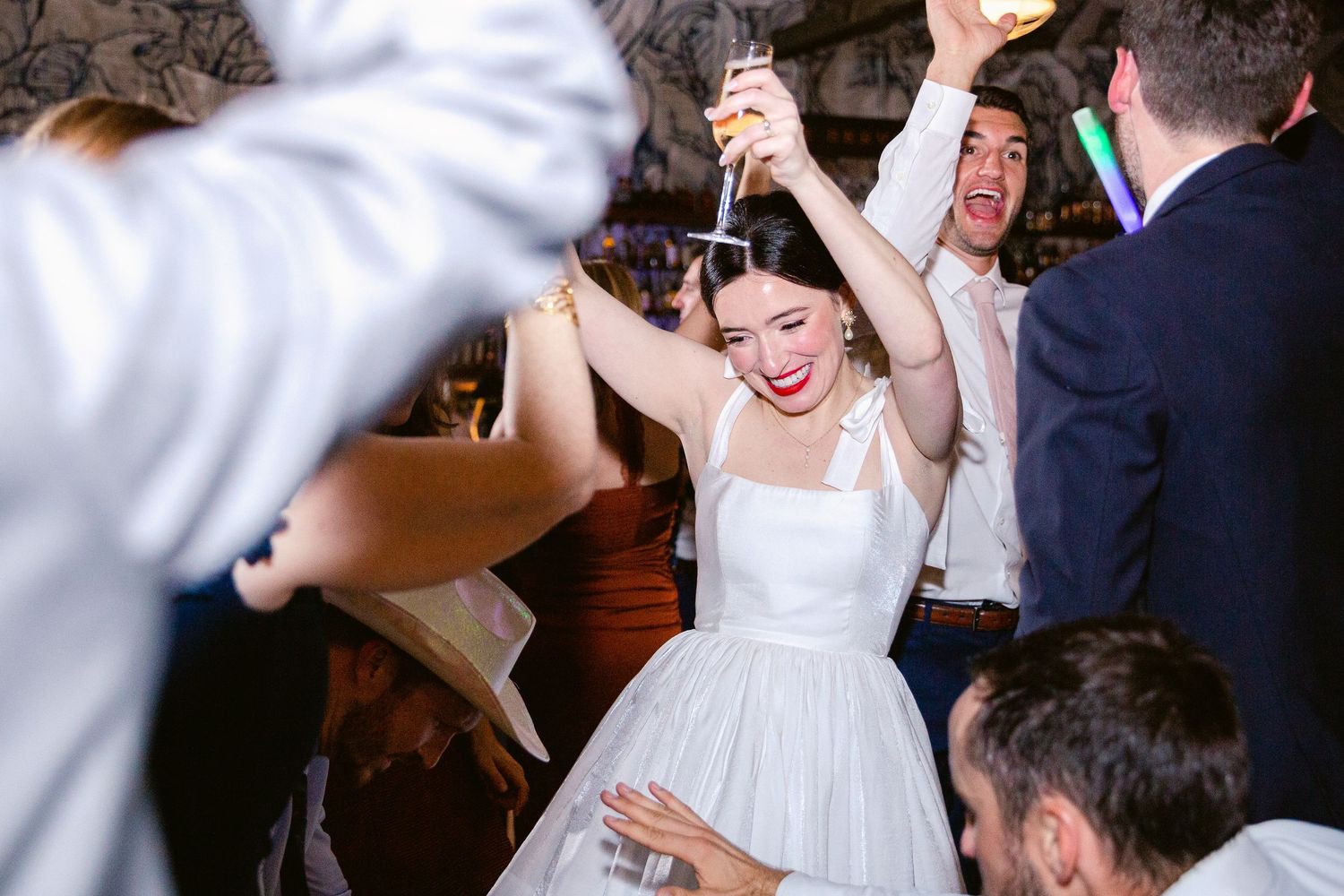 Wedding party dancing energetically at an indoor reception with decorative lighting.