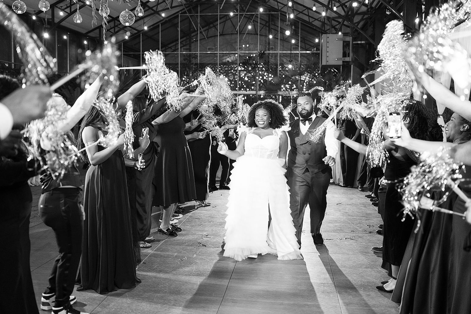 Wedding guests line a pathway holding sparklers as a couple exits their reception in black and white photography.