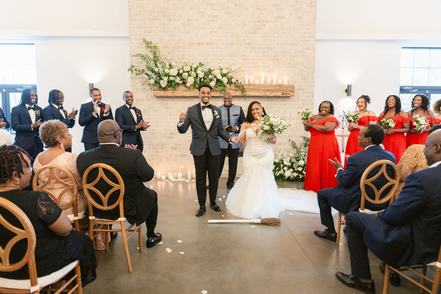 Couple celebrates with wedding party in modern venue with floral decorations and stylish wooden chairs.