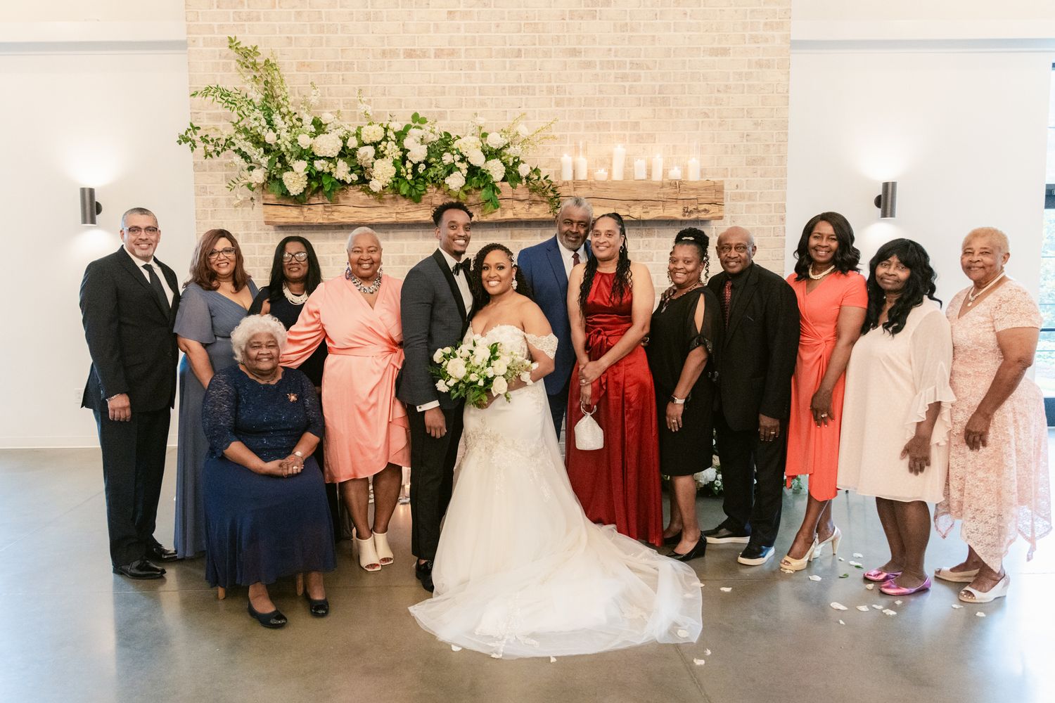 A panoramic wedding party photo showing guests and wedding couple arranged in formal poses against a light colored wall.