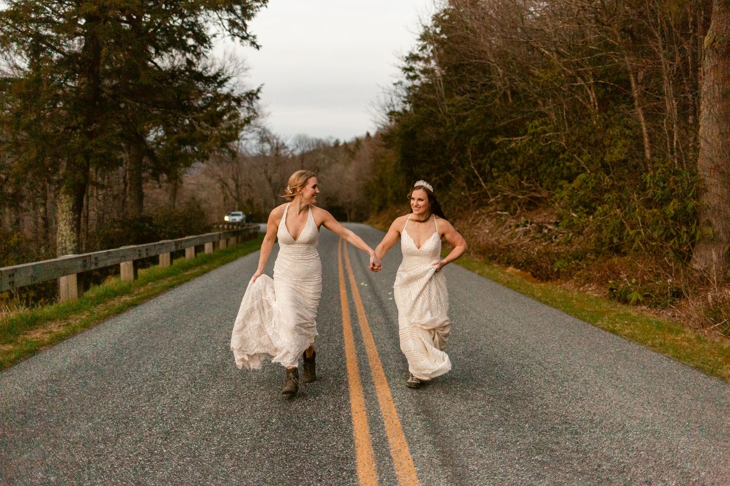 Two brides in white gowns joyfully run hand-in-hand down a scenic tree-lined road on their wedding day.