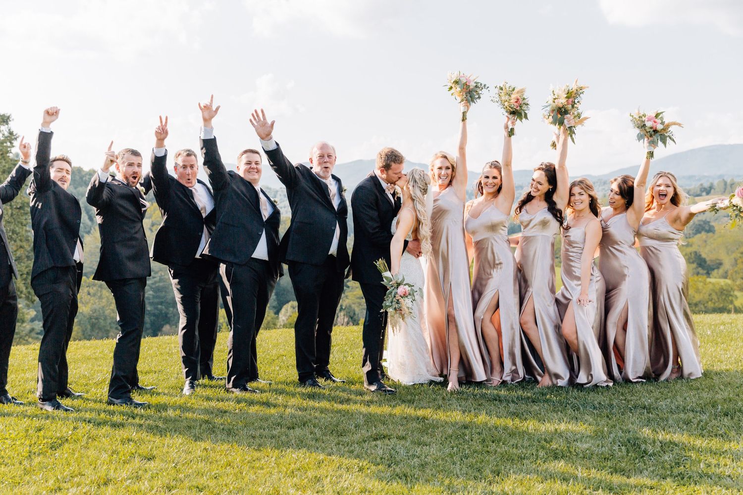 Wedding party jumps in celebration on grassy hill with mountains in background.