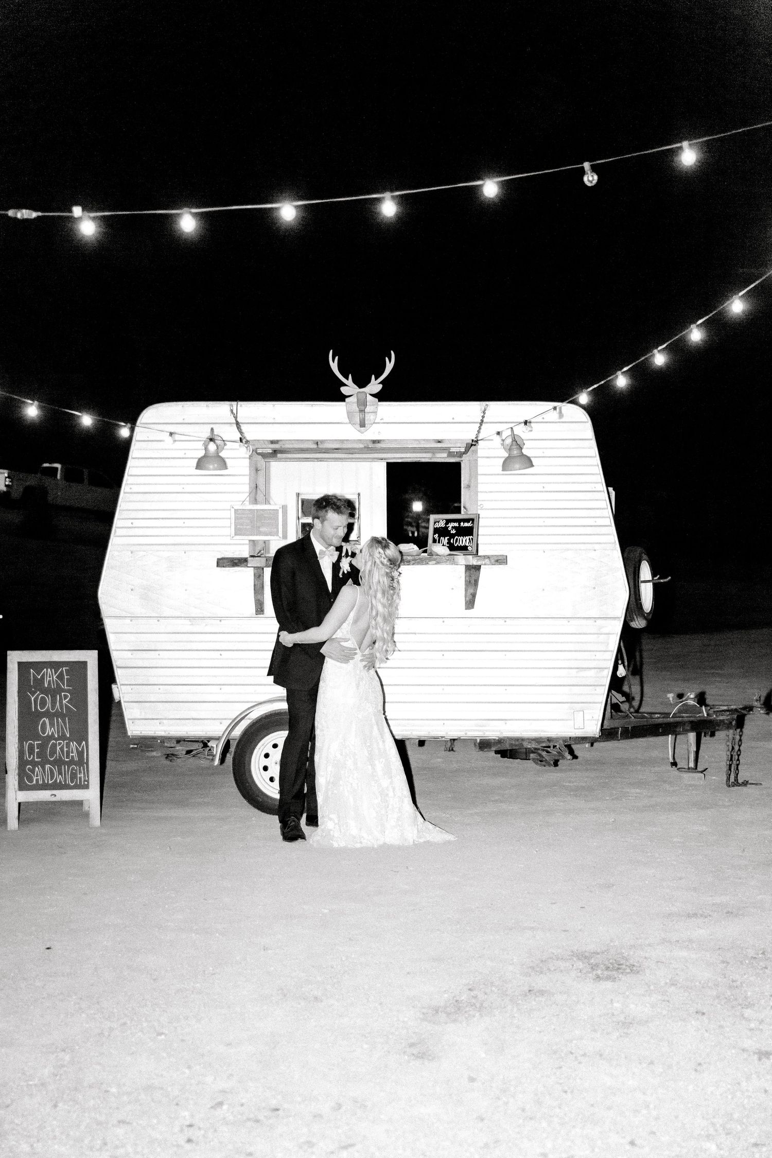 Black and white photo of newlyweds in front of vintage camper trailer with string lights overhead.
