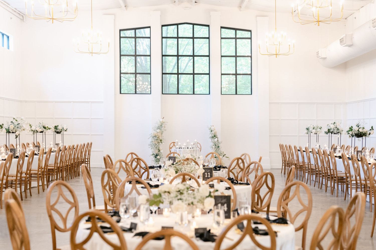 Elegant wedding reception venue with wooden chairs and white decor features tall windows and floral centerpieces.