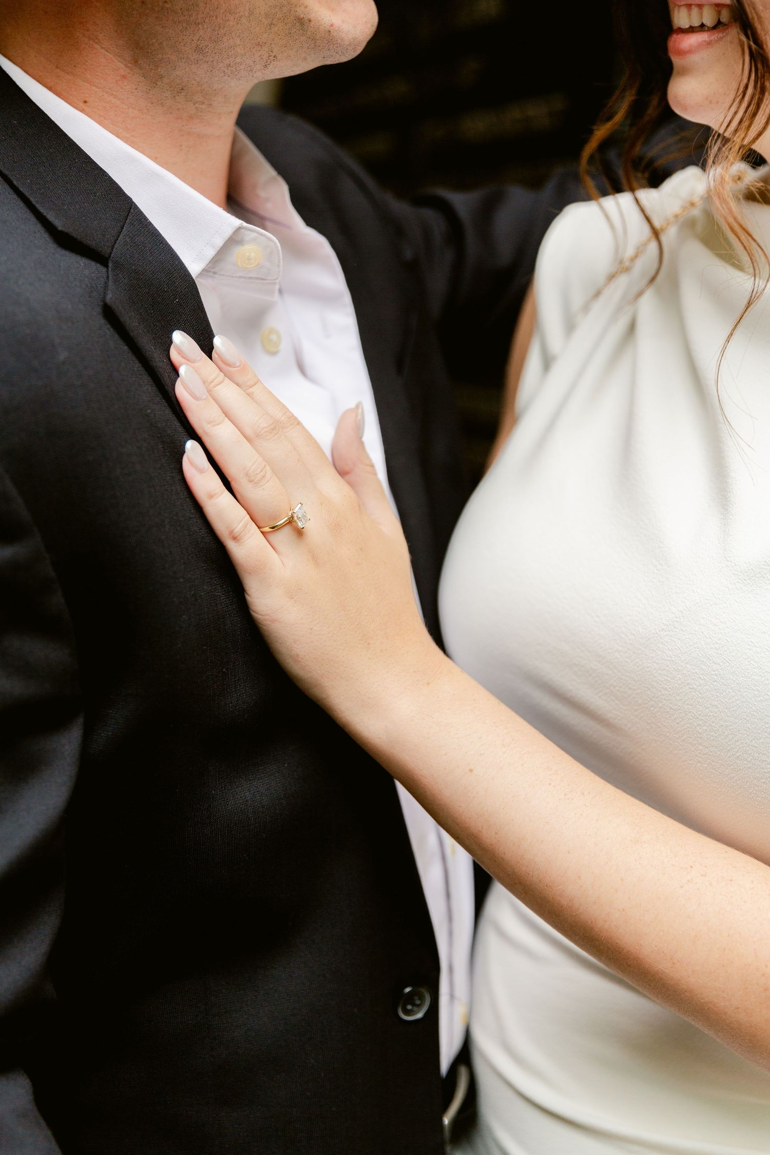 Close-up of bride in white dress touching groom's black suit jacket, showing her delicate gold wedding ring.