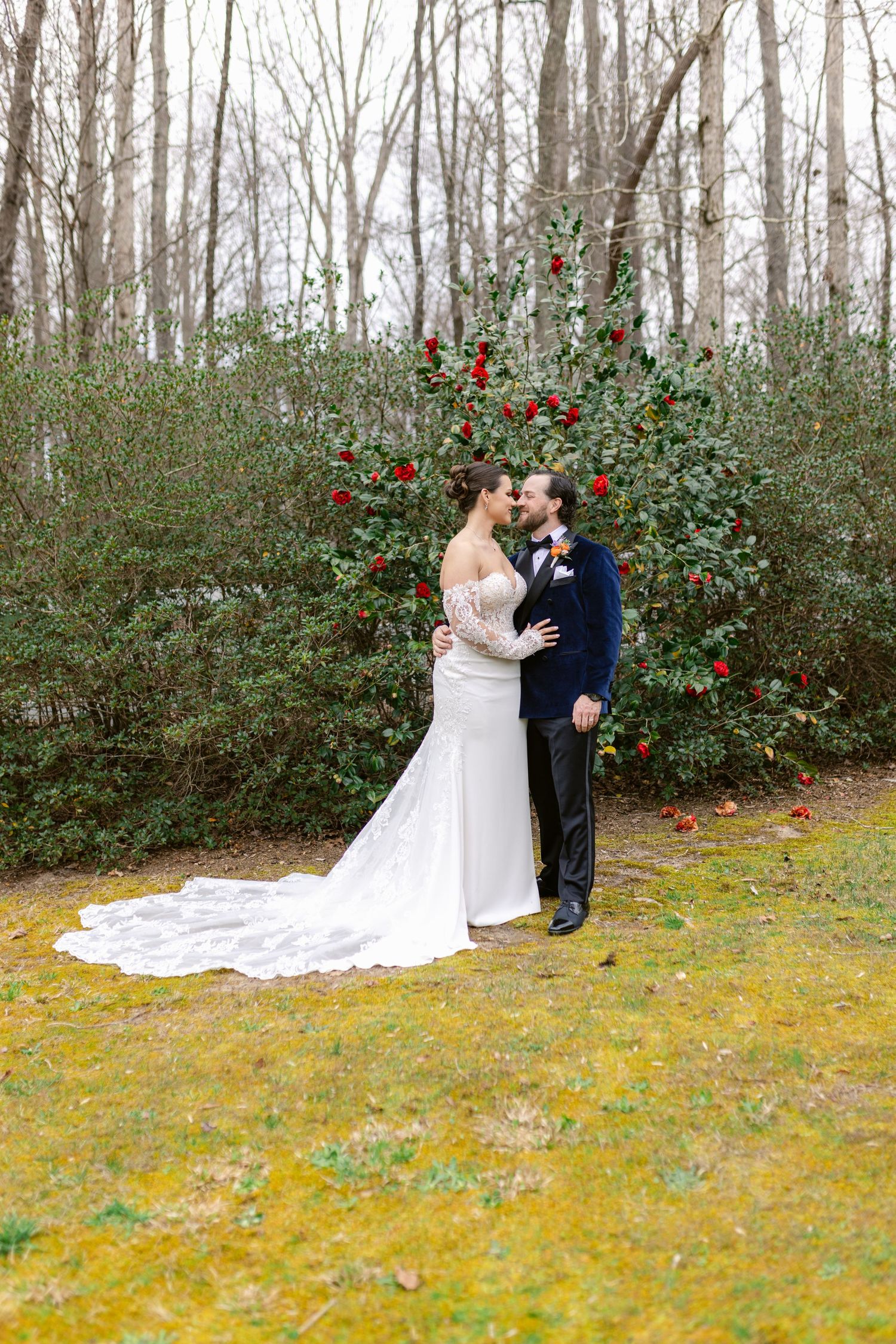 Newlyweds share intimate moment in front of camellia bush on winter day with bride in flowing white gown.
