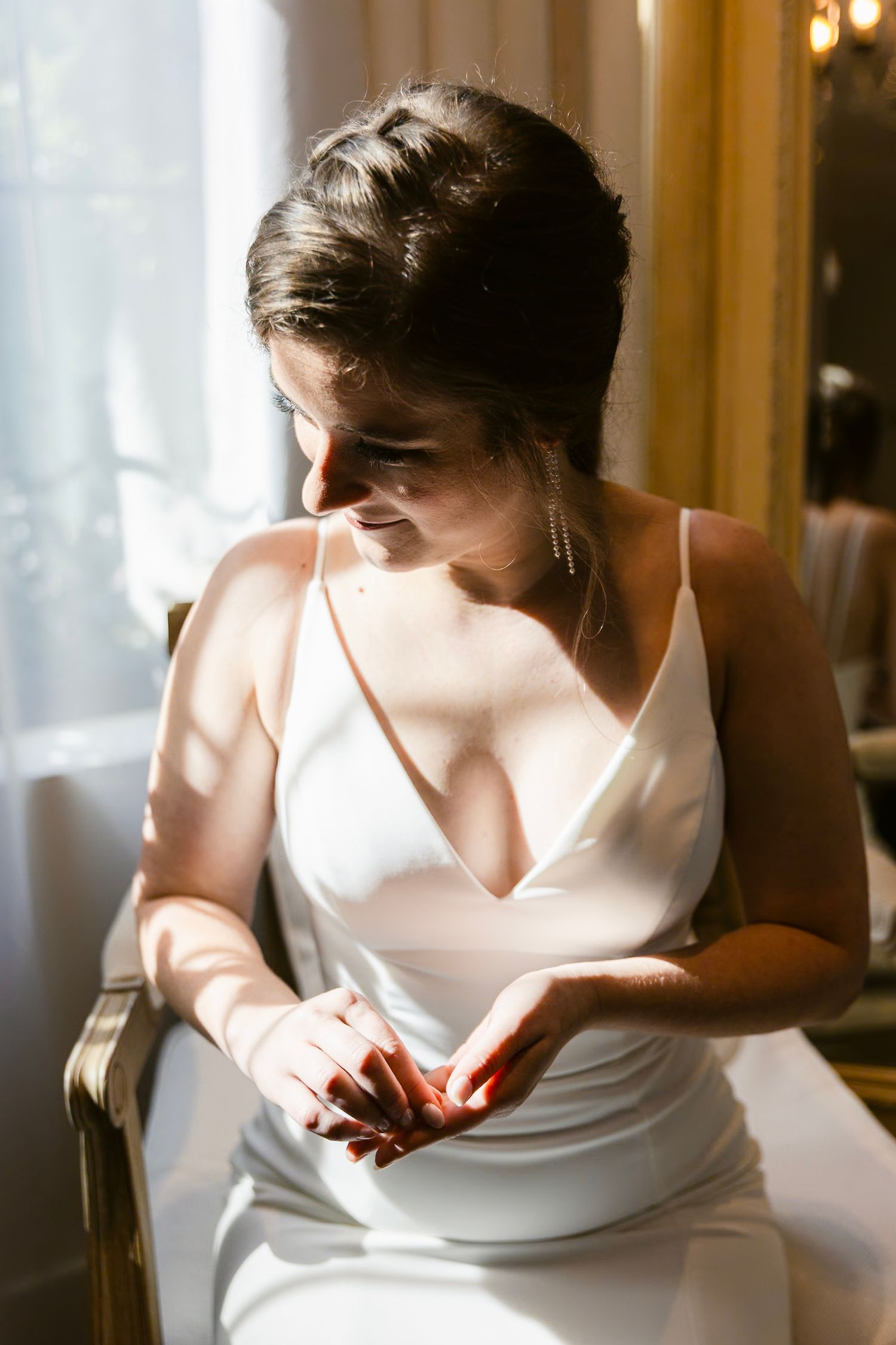 A bride in a simple white satin slip dress sits by a window in soft natural light.