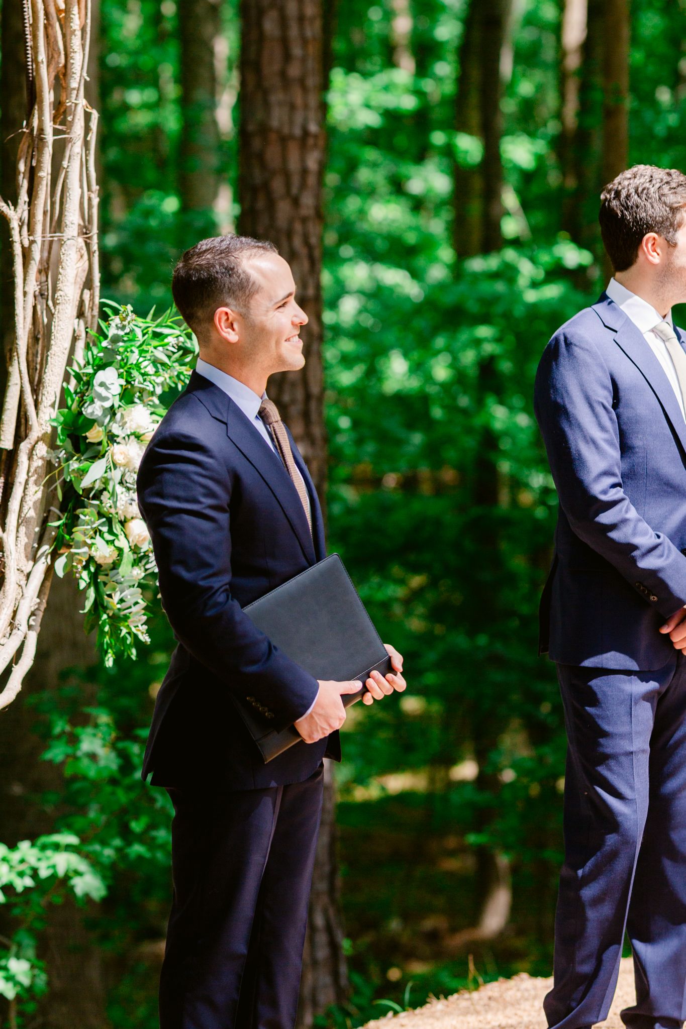 Groomsmen in navy suits stand in a wooded outdoor setting during a wedding ceremony.