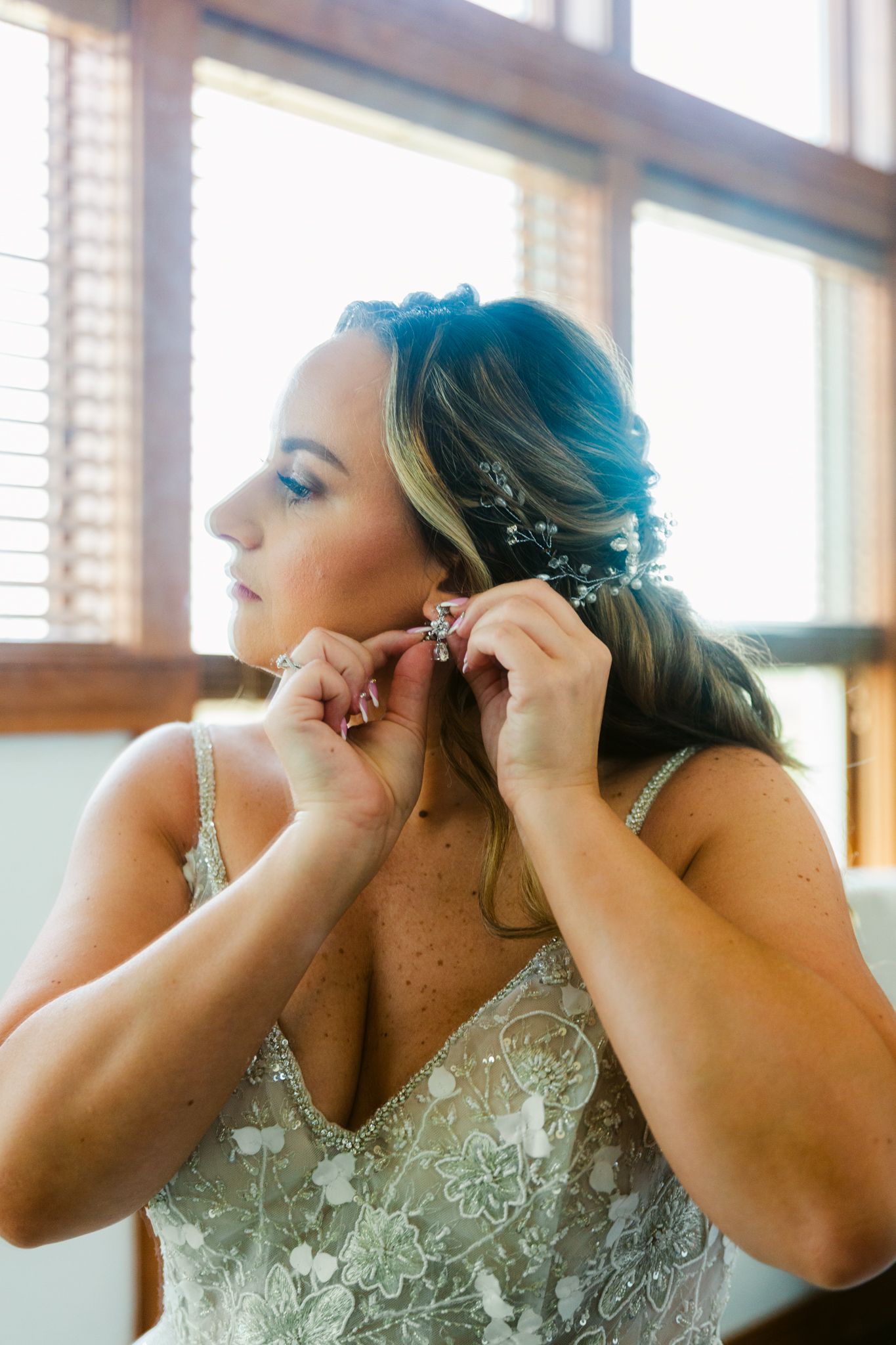 A bride in a beaded lace gown puts on her earrings while getting ready in front of sunlit windows.