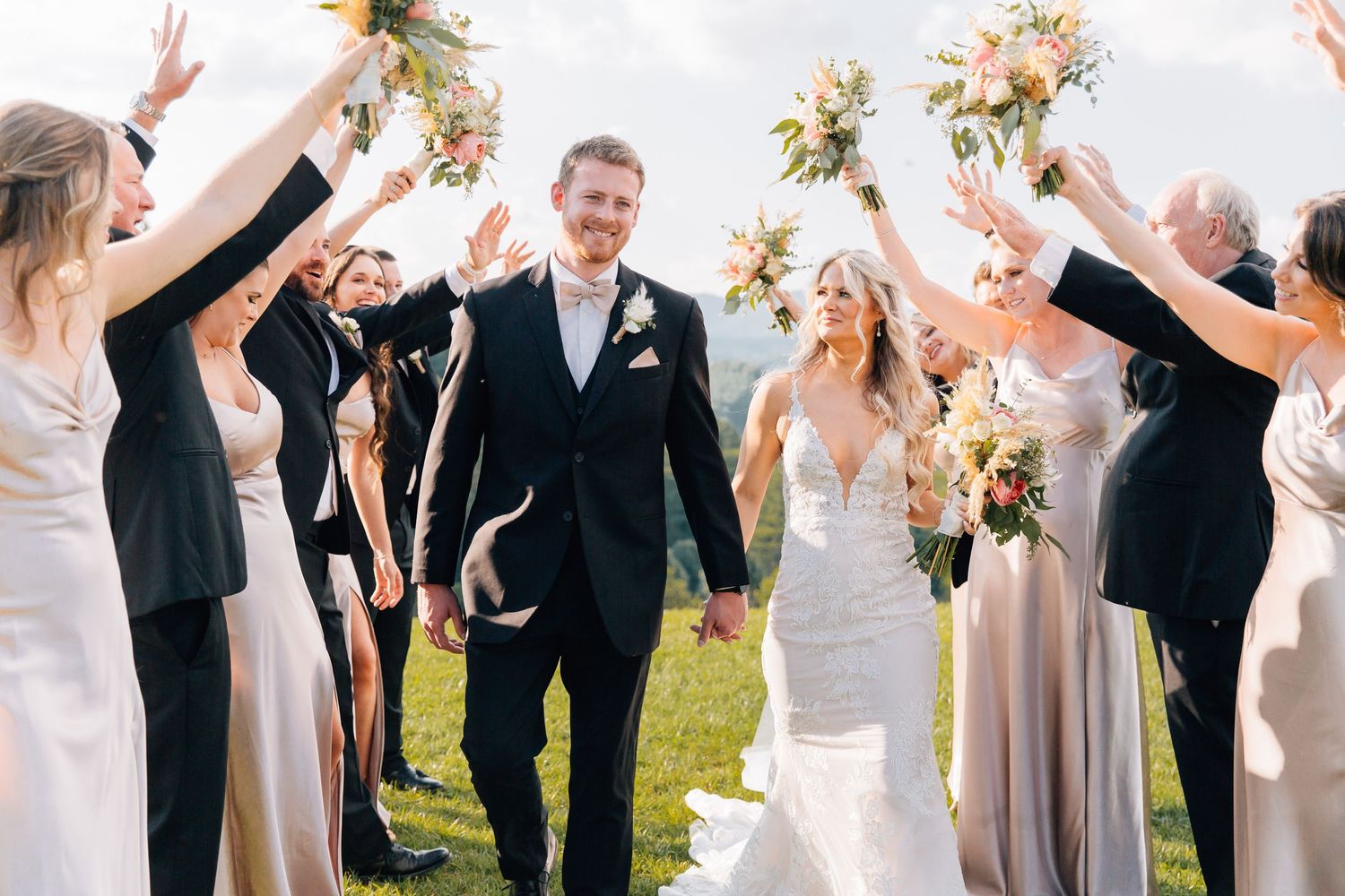 Wedding party in formal attire celebrates joyfully outdoors with raised arms and flower bouquets against scenic backdrop.