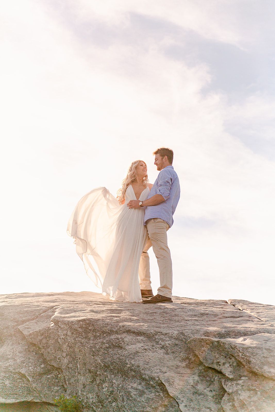 Two people share an intimate moment atop a rocky mountain peak with dramatic clouds in the sky.