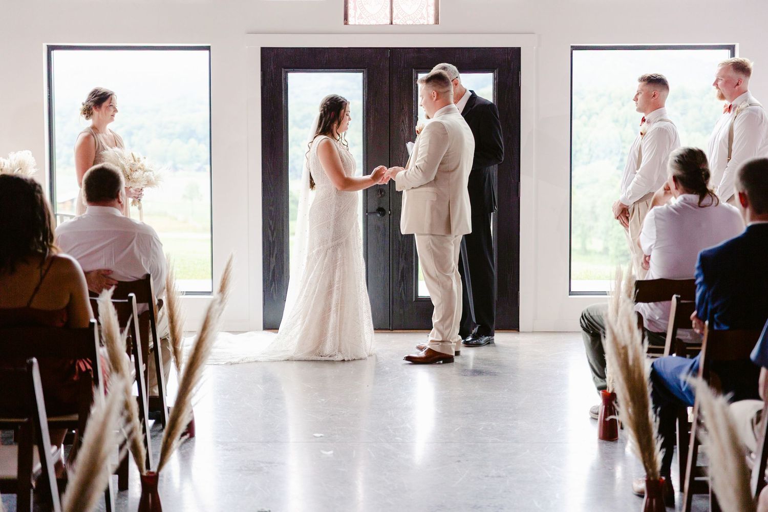 A wedding ceremony takes place in a bright venue with large windows as guests watch the couple exchange vows.