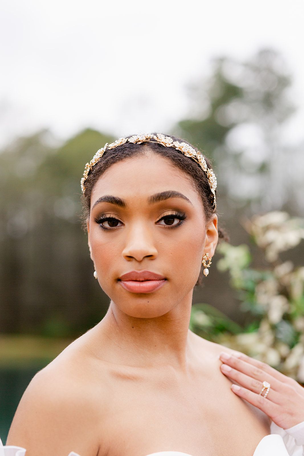 A bridal portrait with delicate gold headpiece, strapless gown and soft natural makeup in an outdoor setting.