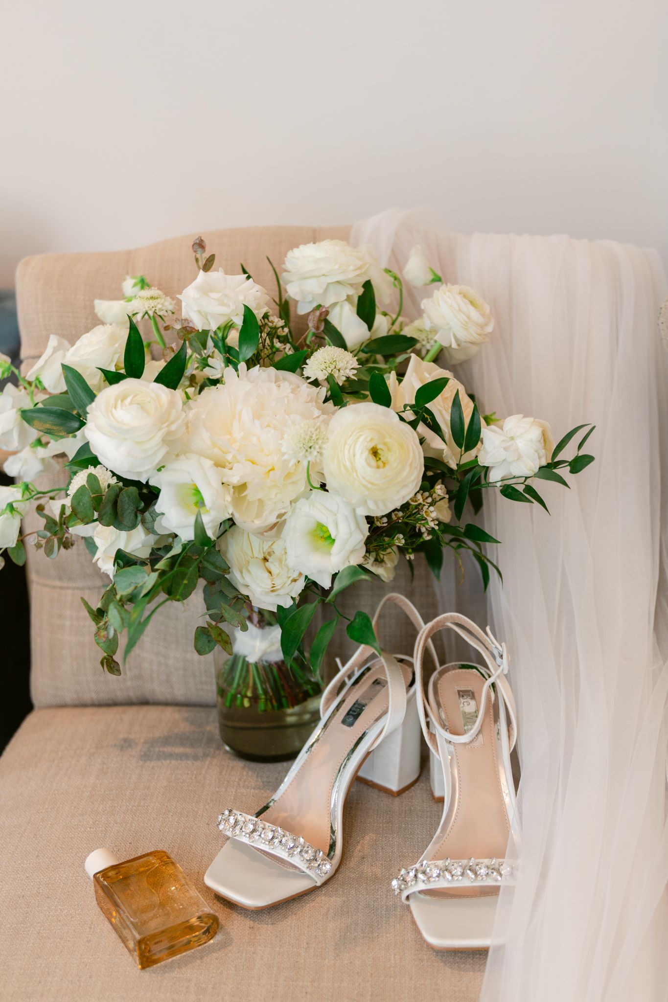 Elegant white floral arrangement with roses and greenery next to bridal heels and perfume bottle on beige chair.