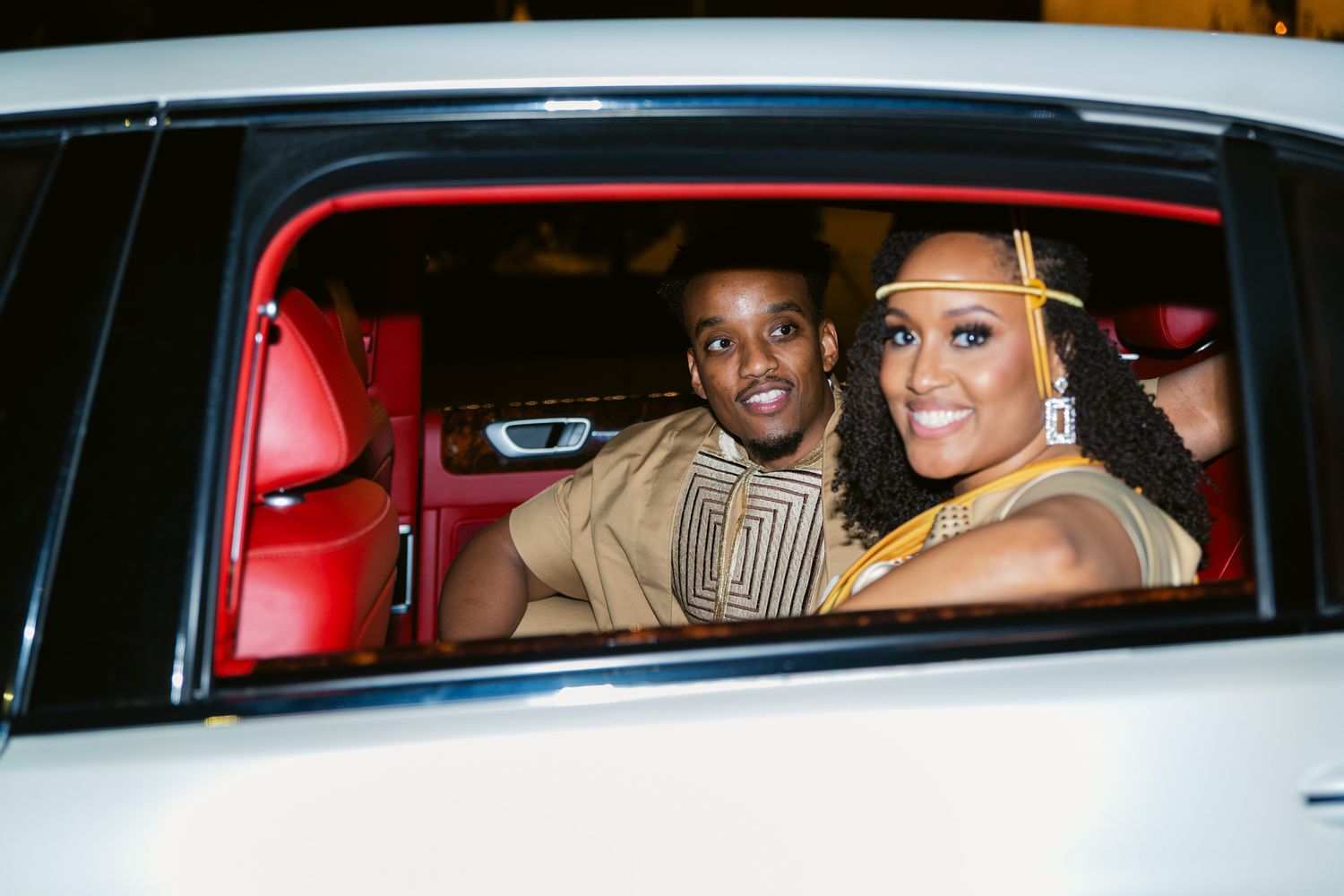 Two people smile while sitting in a luxury car with red leather interior viewed through the passenger window.