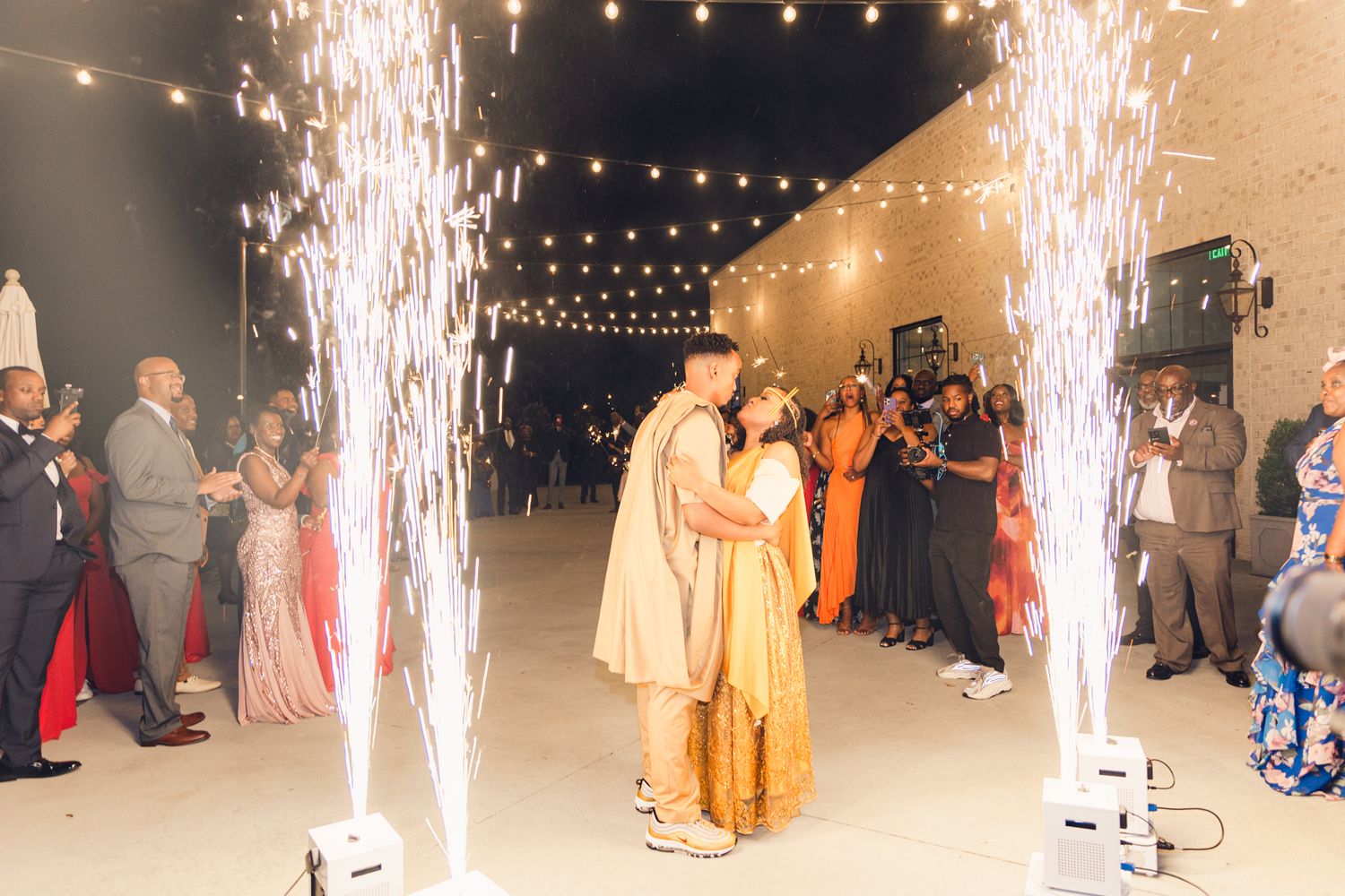 Sparklers create a magical atmosphere at an evening outdoor celebration with string lights and guests in formal attire.