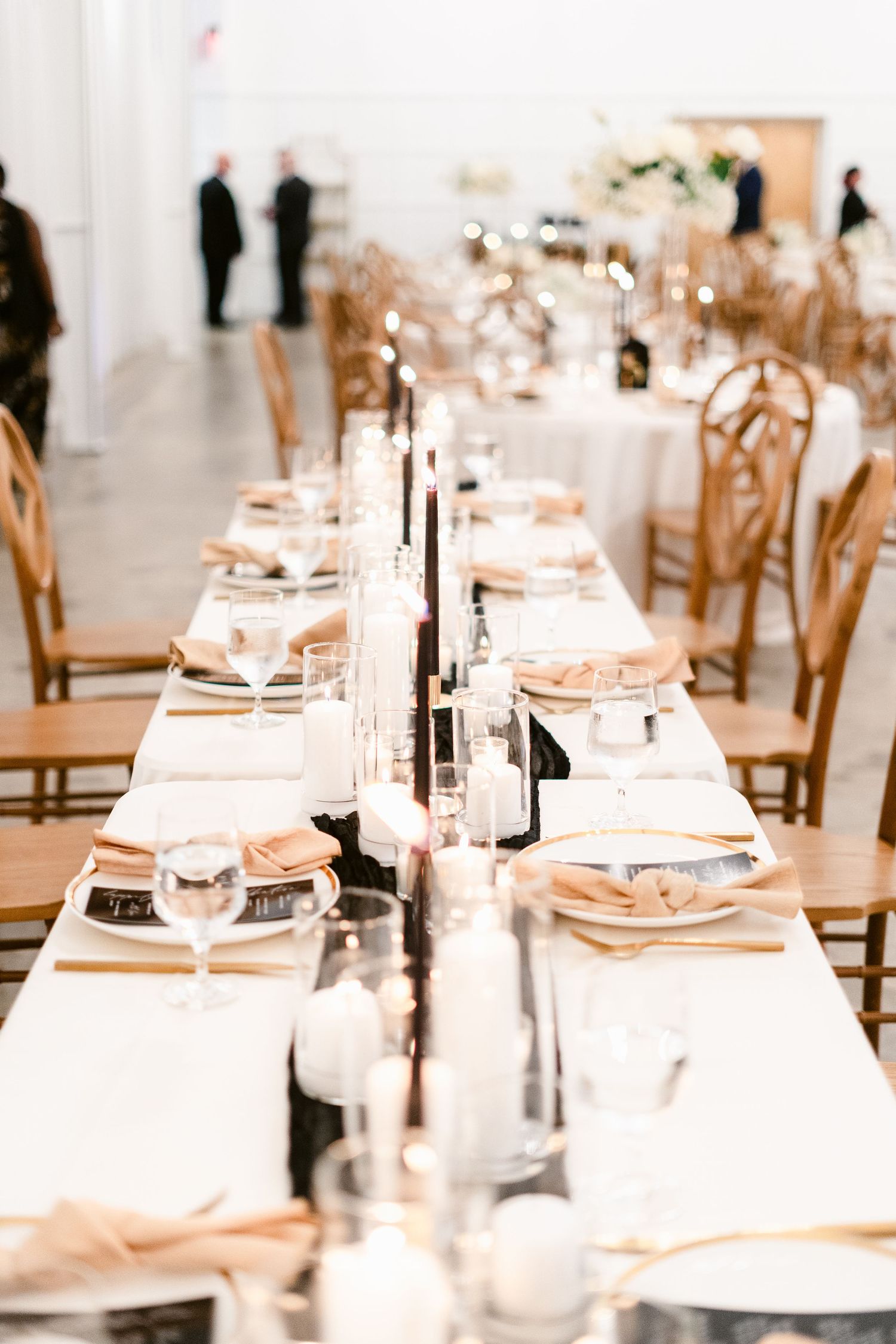 Elegant wedding reception table setting with white linens, tall black candles, and wooden chairs in a bright venue.