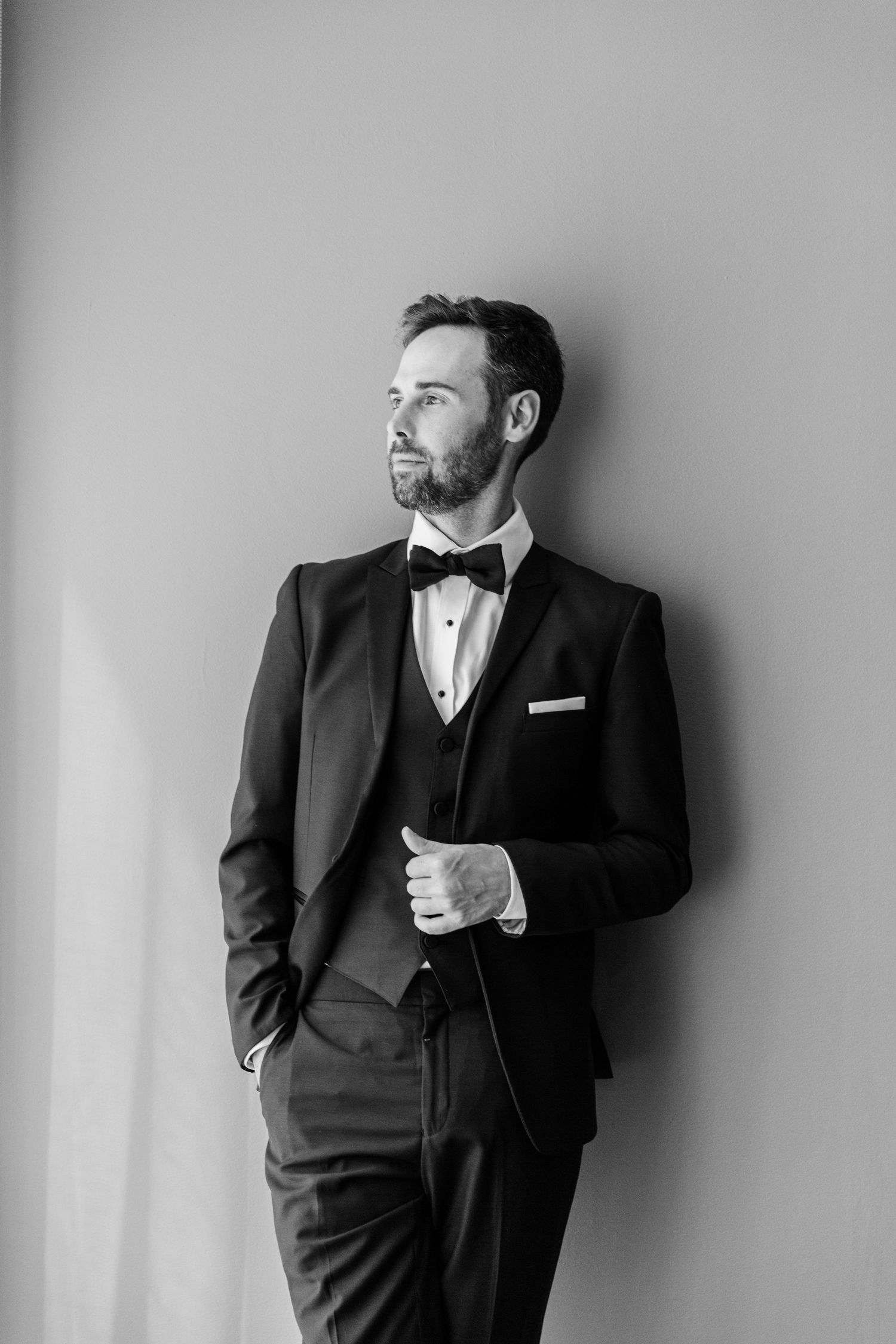 Black and white portrait of a person in formal tuxedo with bow tie looking to the side against a plain wall.