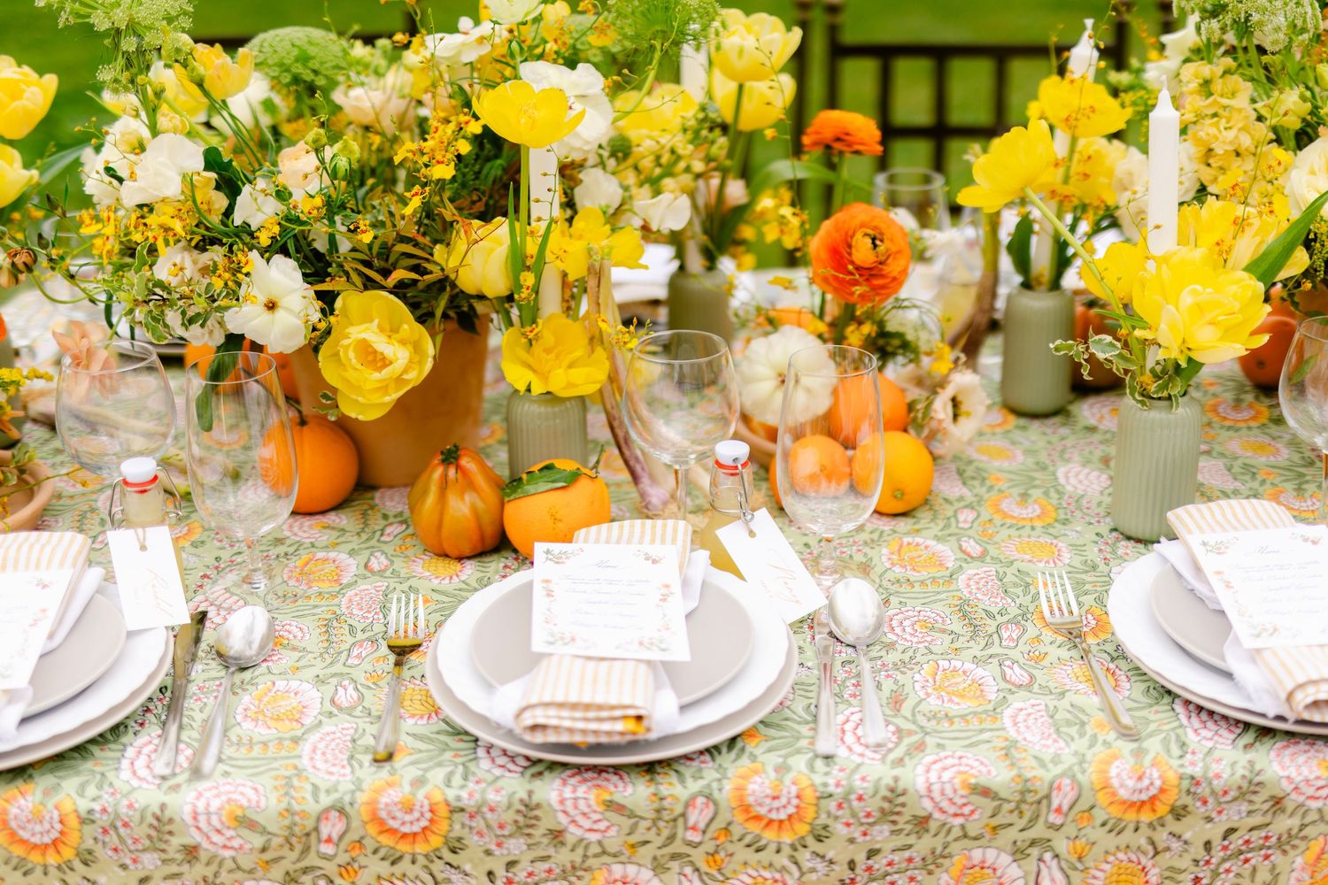 A festive fall table setting with yellow and orange flowers, pumpkins, and elegant place settings on a floral tablecloth.