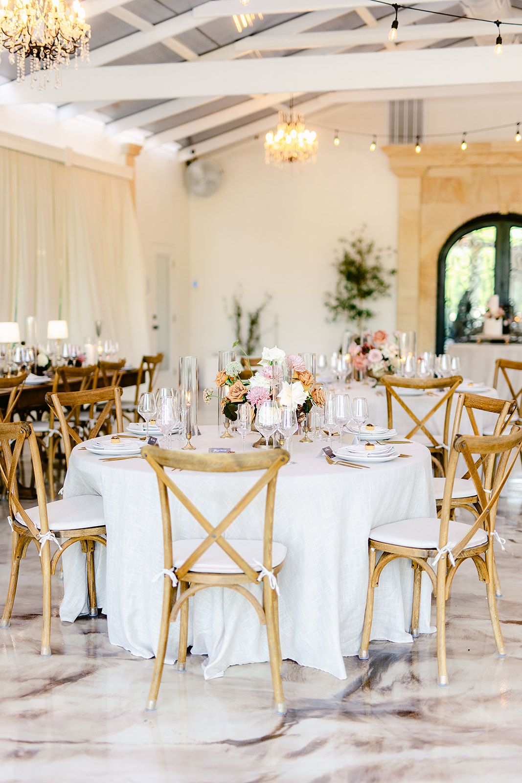 Light-filled banquet hall with crystal chandeliers, round dining tables, and rustic wooden chairs set for an event.