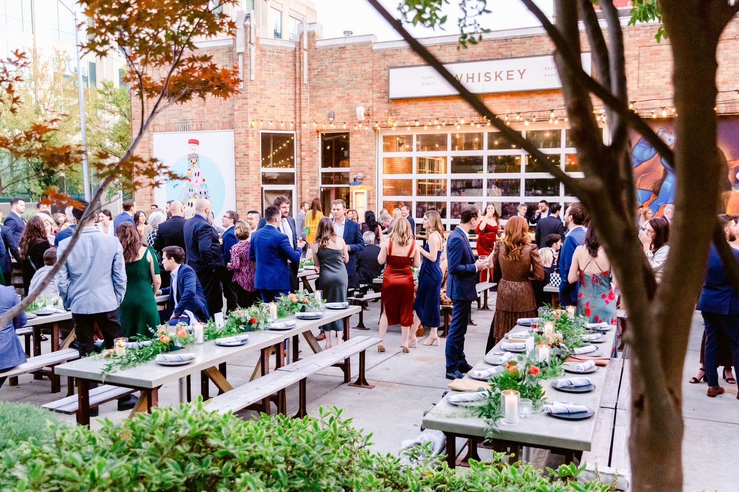 Outdoor wedding reception setup with long picnic tables and string lights against brick building backdrop.