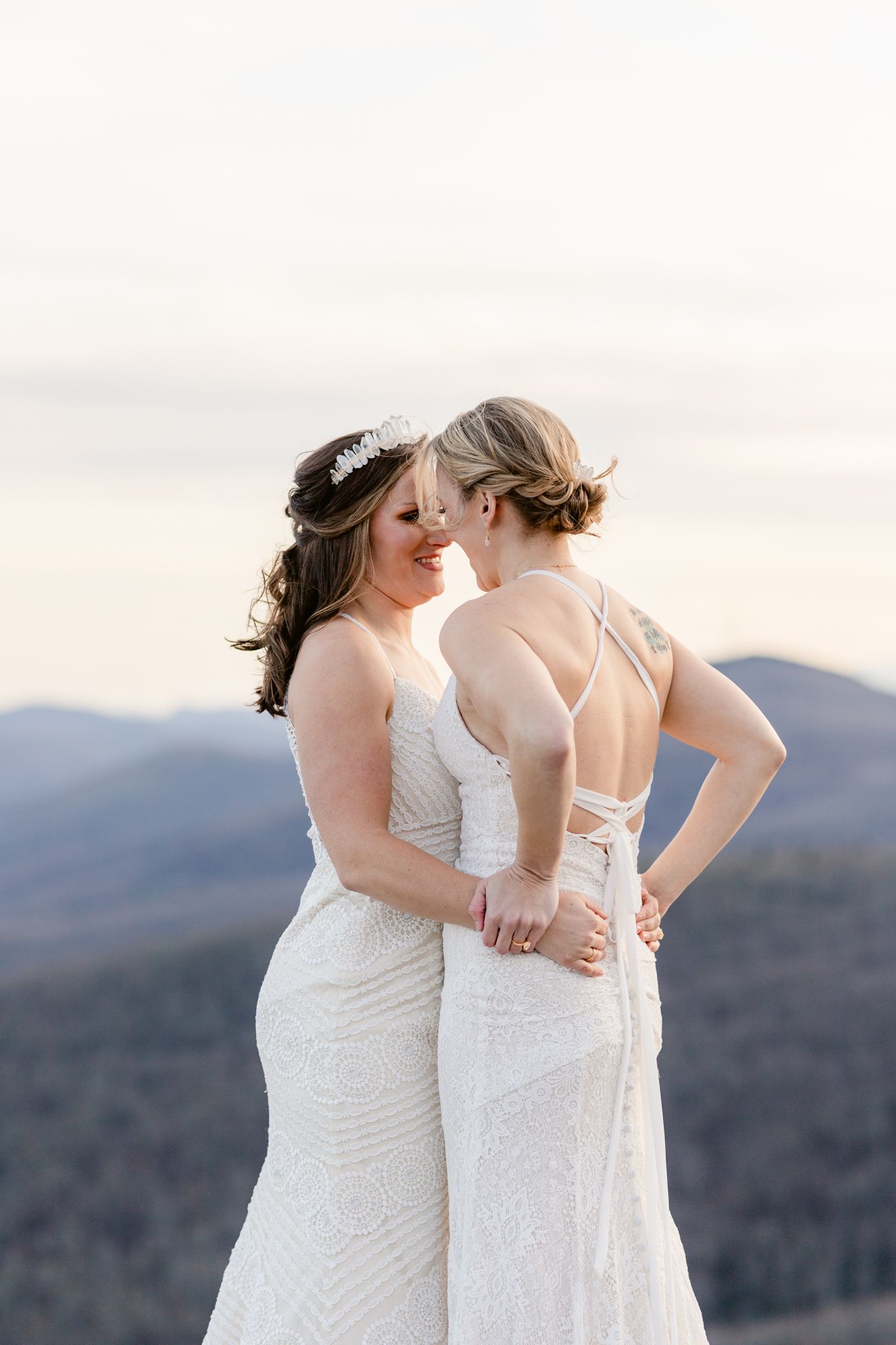 Two brides in white wedding dresses share an intimate moment against a mountain backdrop at sunset.
