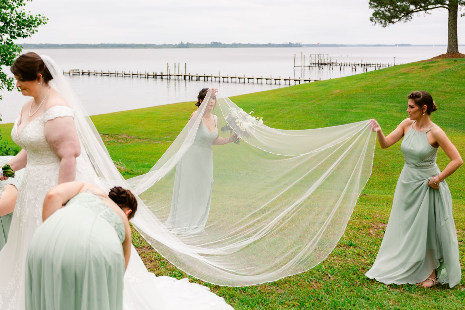 Wedding party members hold a flowing white veil on a grassy lawn overlooking a waterfront pier.