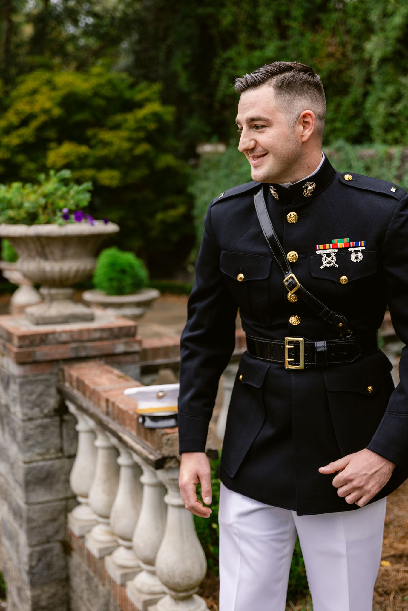 Marine in dress uniform leans against stone balustrade in formal garden setting during photo session.