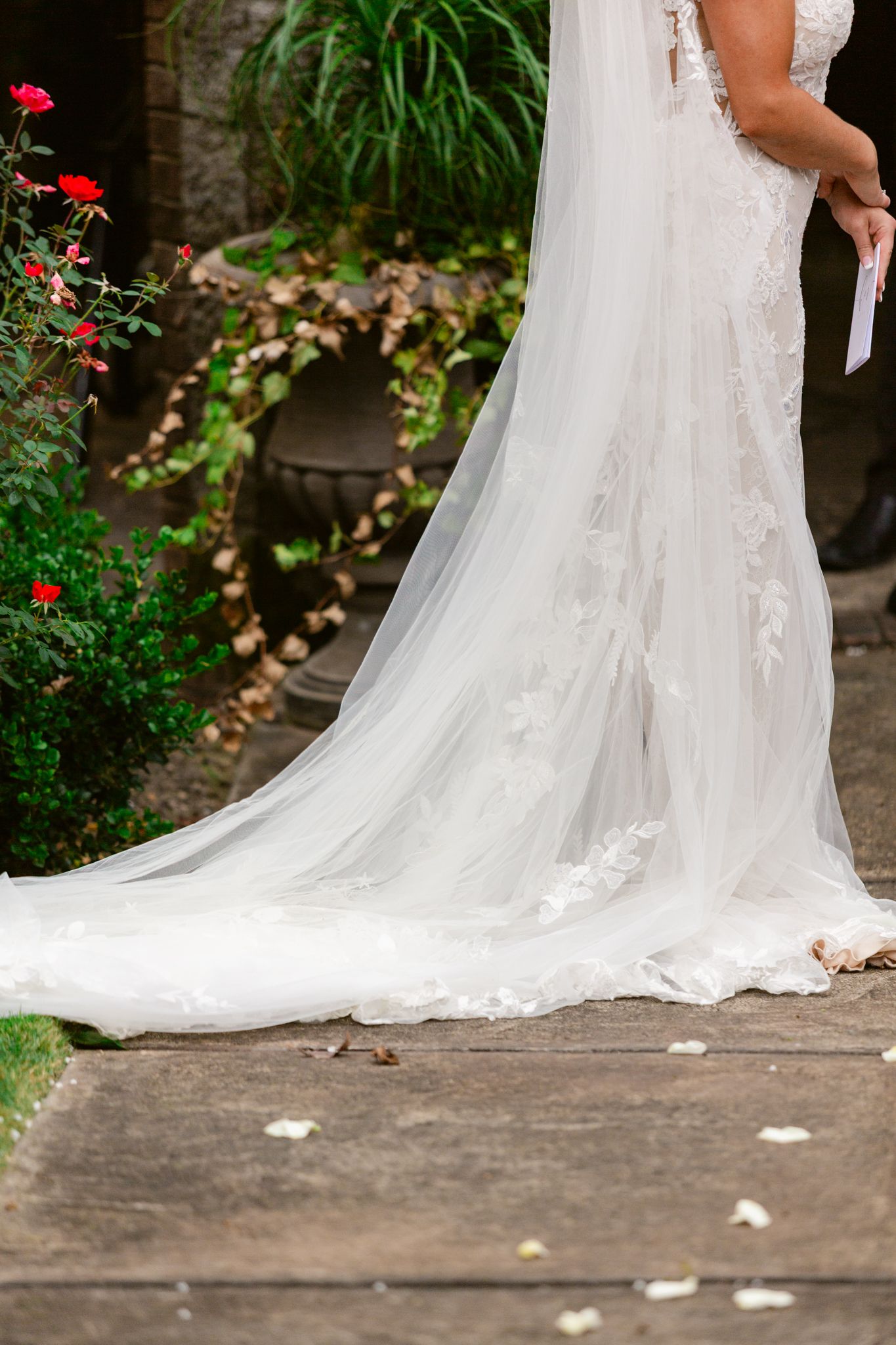 A flowing white wedding dress train with delicate lace details trails across a stone patio with red roses in the background.