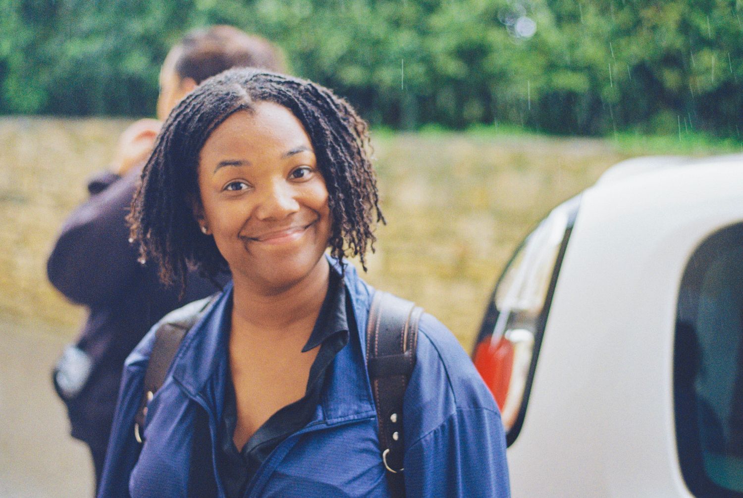 A person with braided hair wearing a navy jacket smiles warmly while standing next to a parked car outdoors.