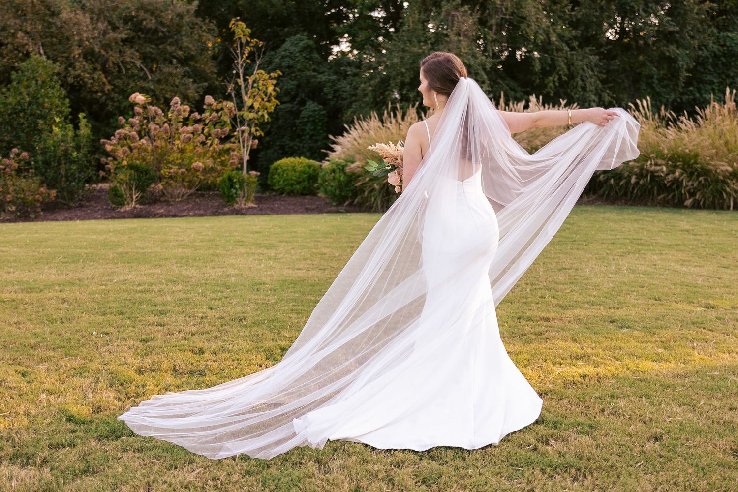 Flowing white wedding dress and long veil billowing in the wind on a garden lawn with autumn flowers in background.