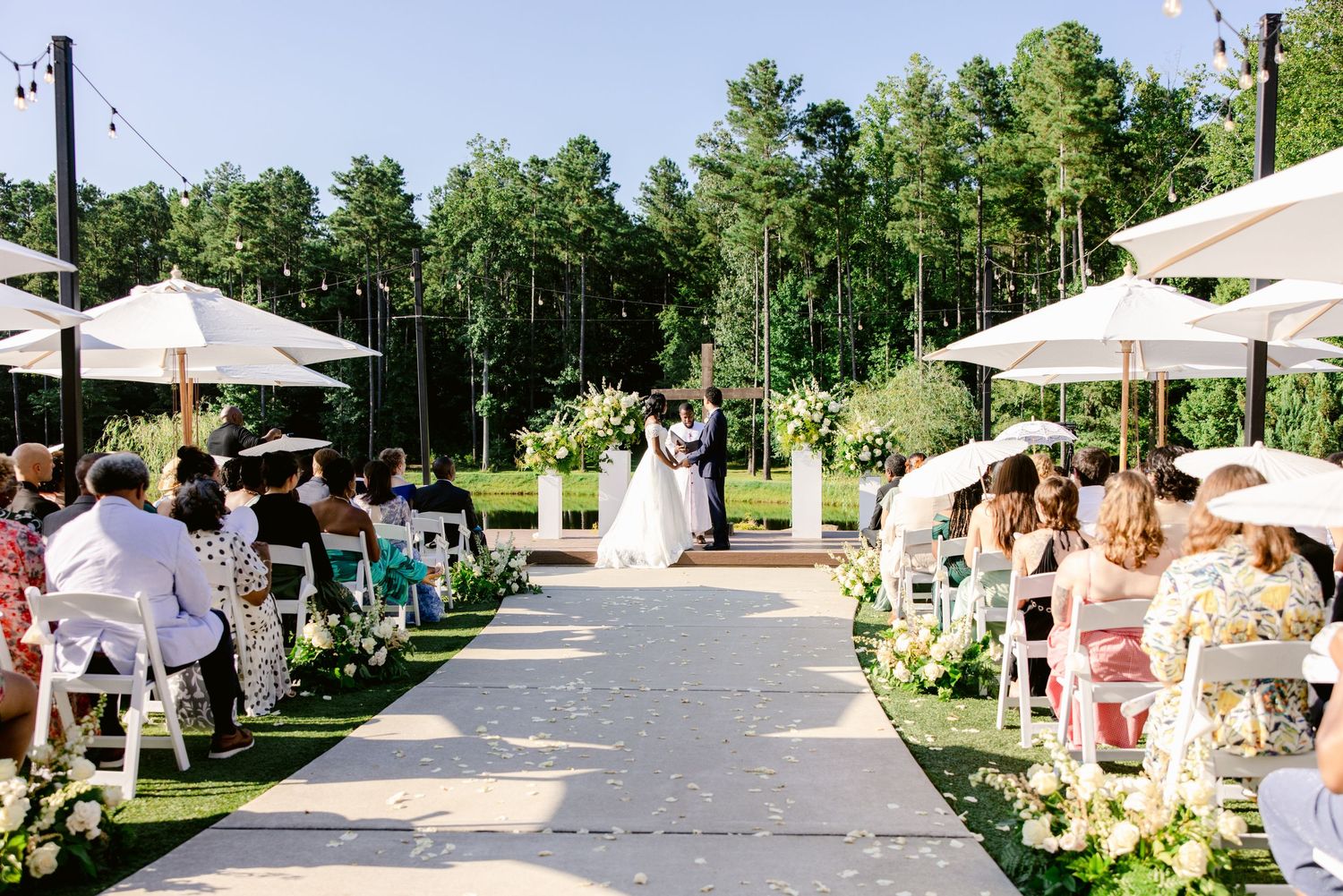 Outdoor wedding ceremony on a sunny day with white umbrellas and guests seated on white chairs along a stone pathway.