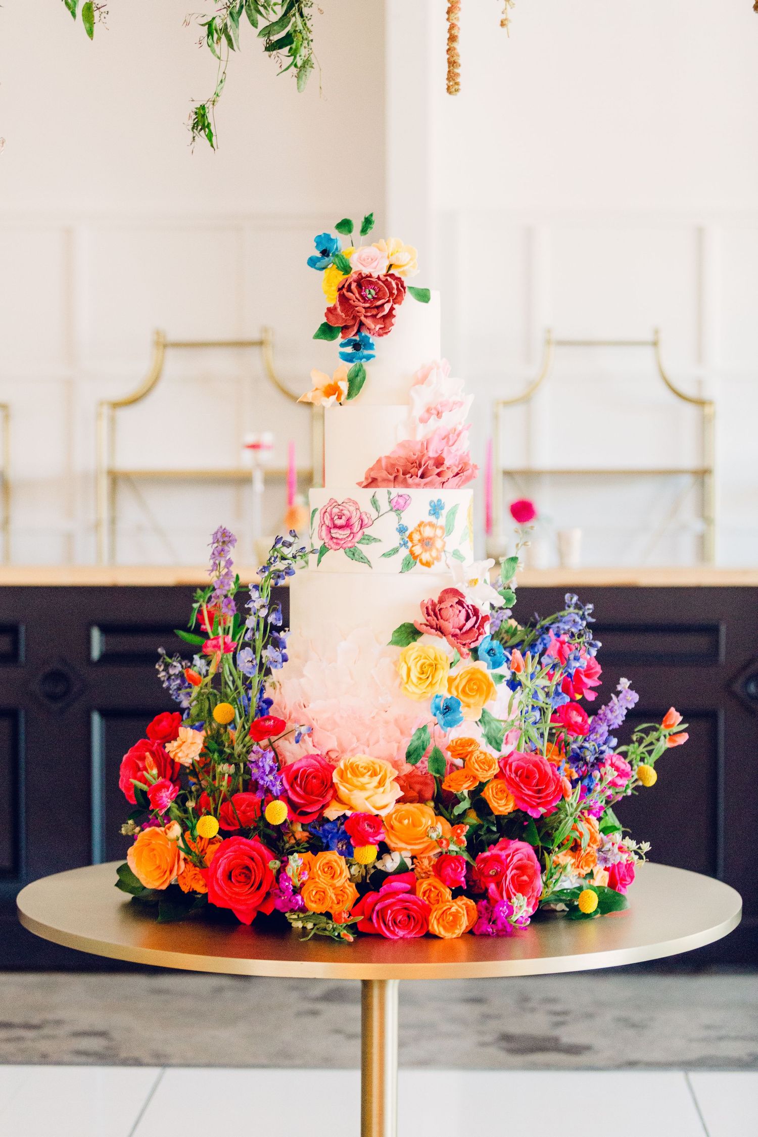 A white wedding cake adorned with vibrant coral, yellow and blue fresh flowers arranged in a cascading pattern.