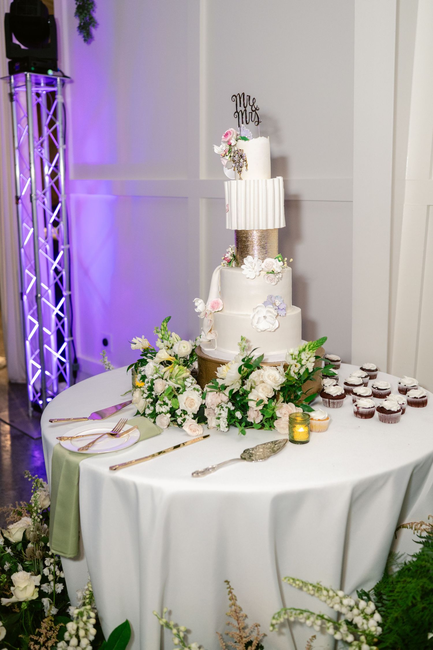 Elegant wedding cake display table with white linens, purple uplighting, and floral arrangements featuring white blooms and greenery.