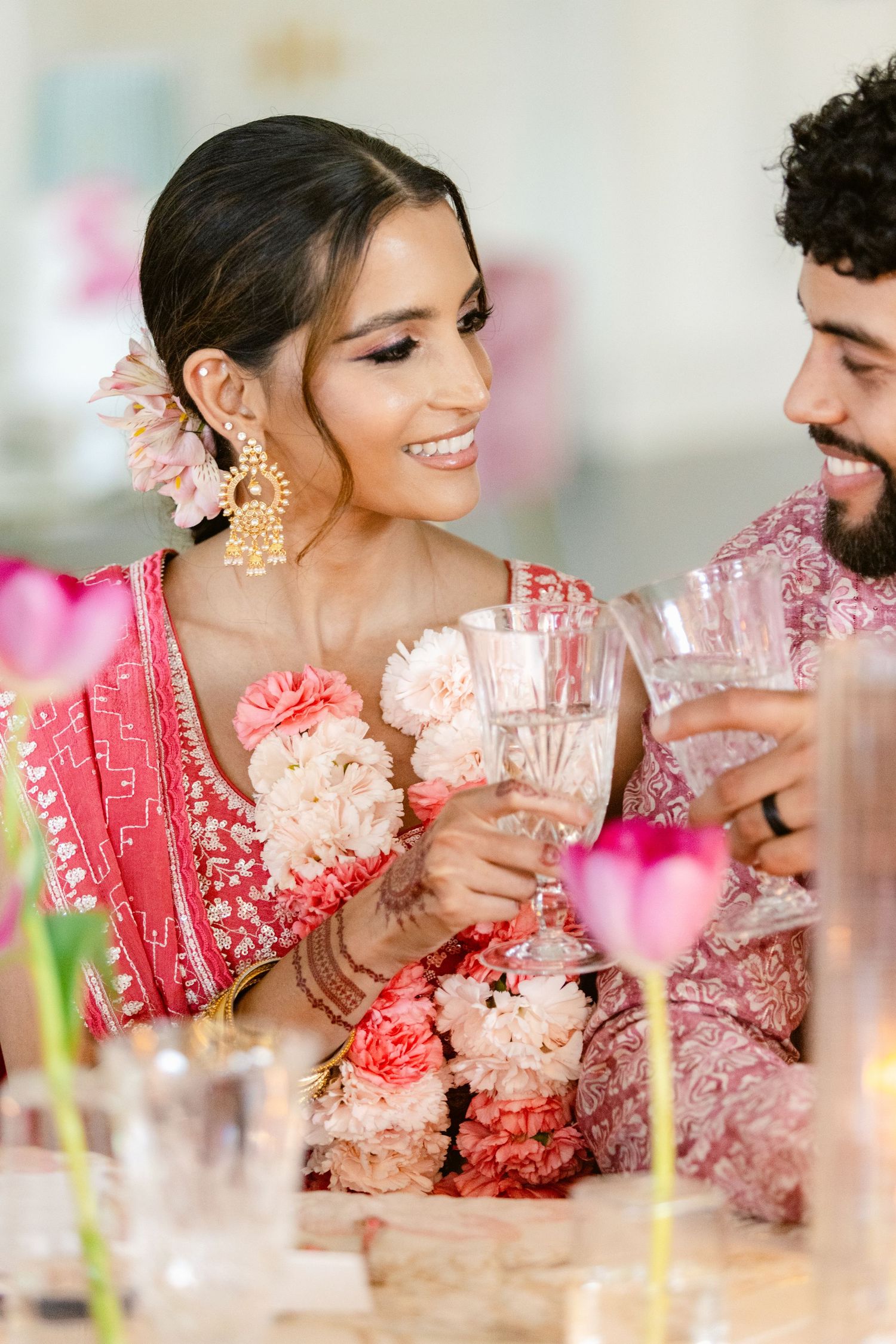 A couple in traditional Indian wedding attire share a tender moment among pink flowers and soft lighting.