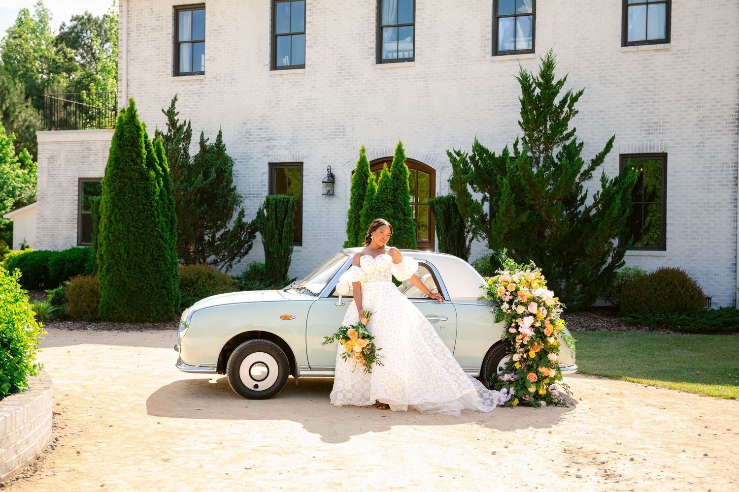 A vintage silver car decorated with flowers parked in front of a white stucco building with cypress trees.