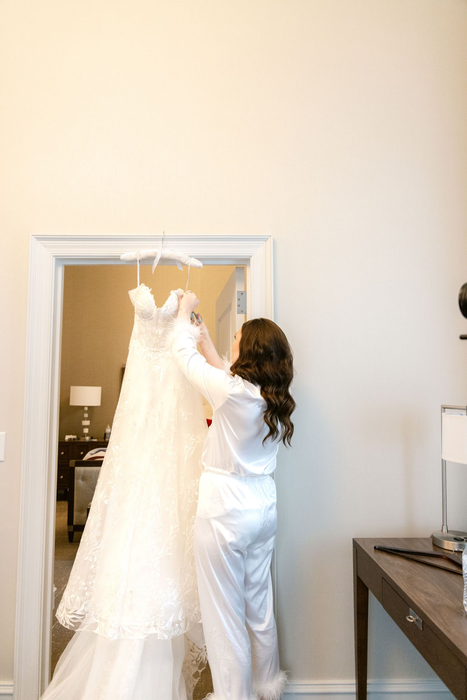A person in white silk pajamas hangs up an elegant lace wedding dress in a brightly lit room.