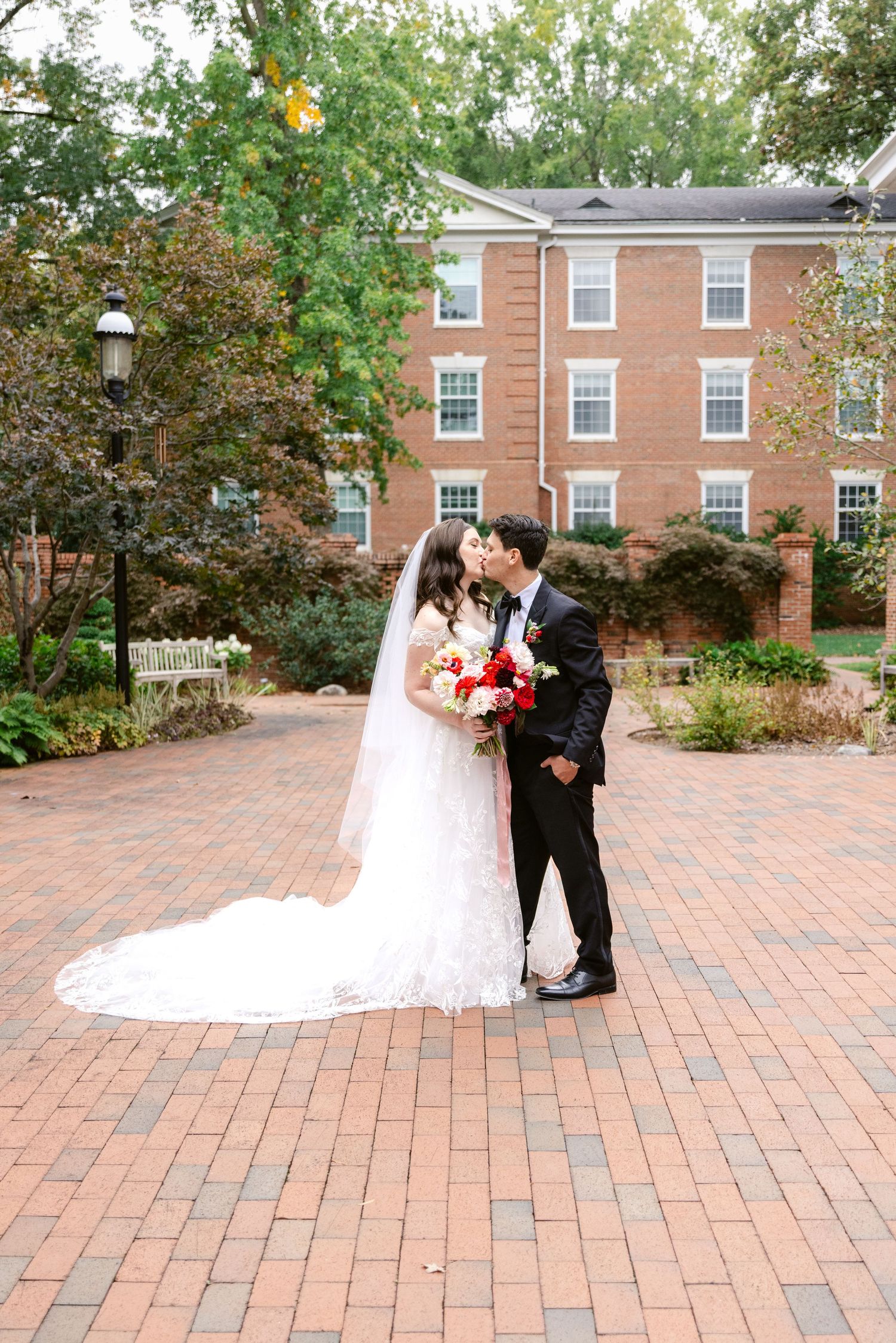 A romantic wedding moment captured on a brick courtyard with fall foliage and brick buildings in the background.