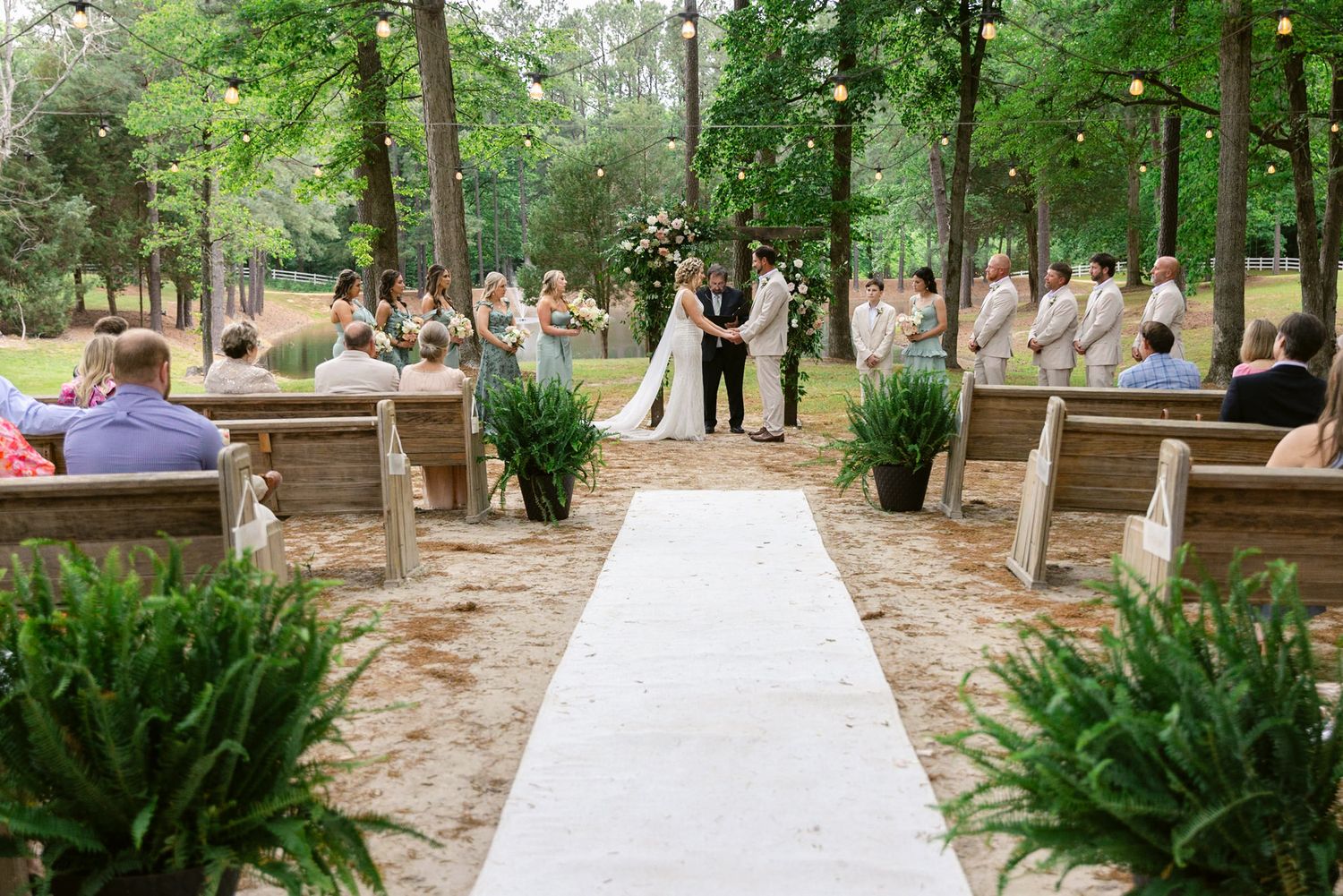 A beautiful outdoor wedding ceremony takes place in a wooded setting with guests seated on rustic wooden benches.