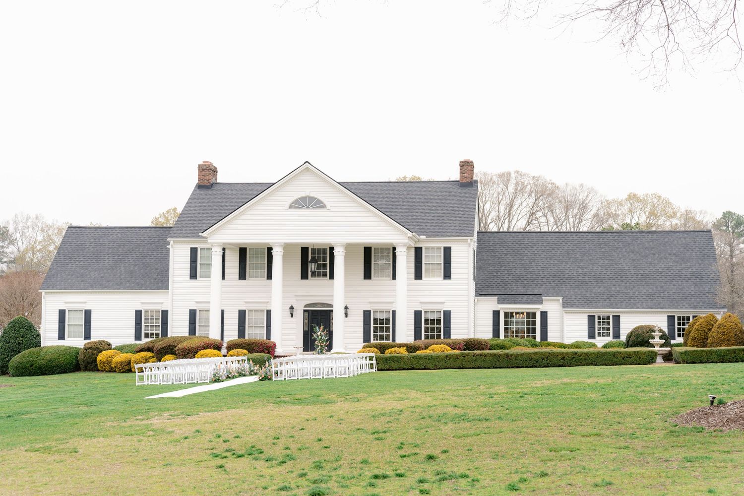 White colonial style house with black shutters sits on a spacious green lawn with mature trees in the background.