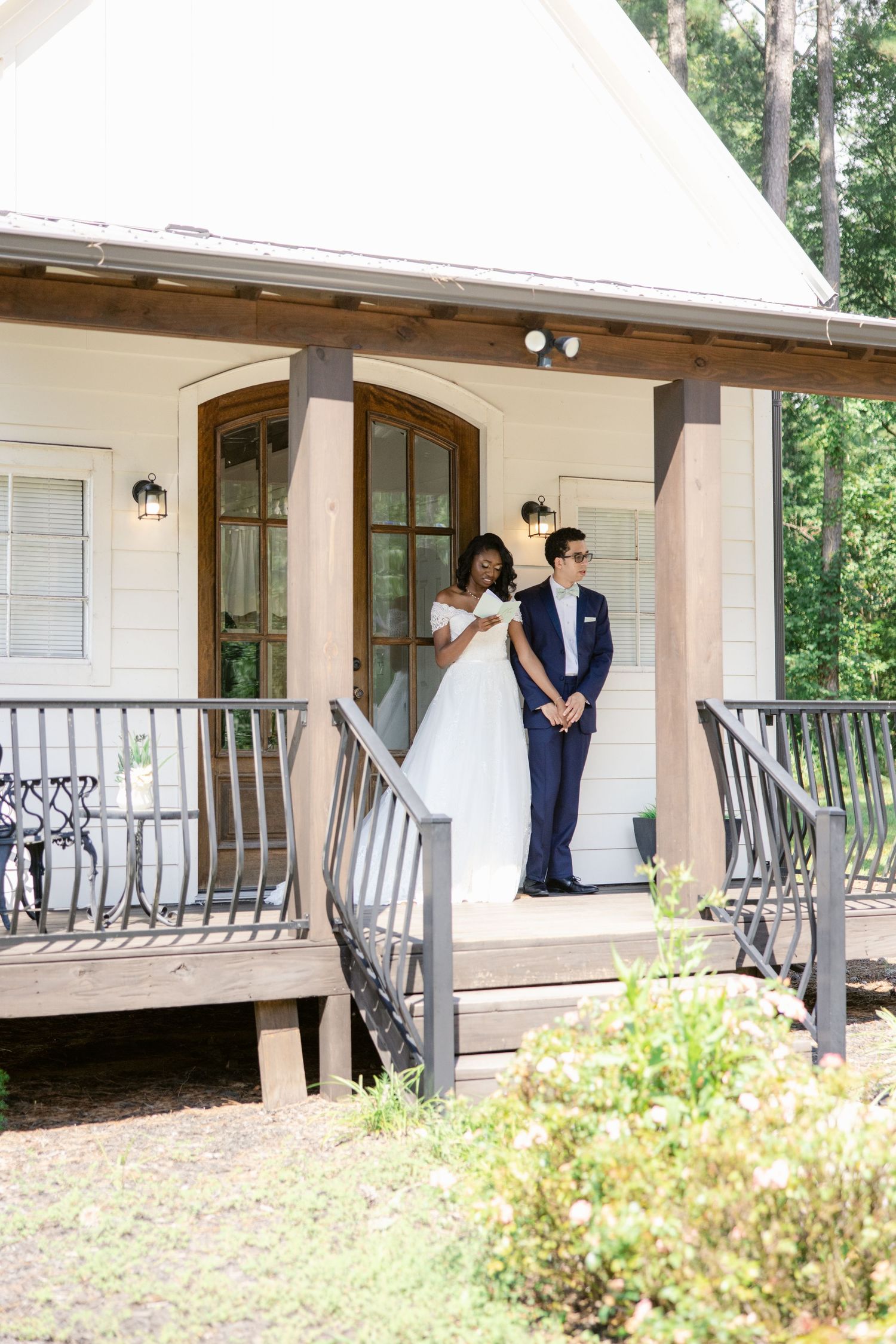 A couple in formal wedding attire share an intimate moment on a rustic covered porch with white railings.