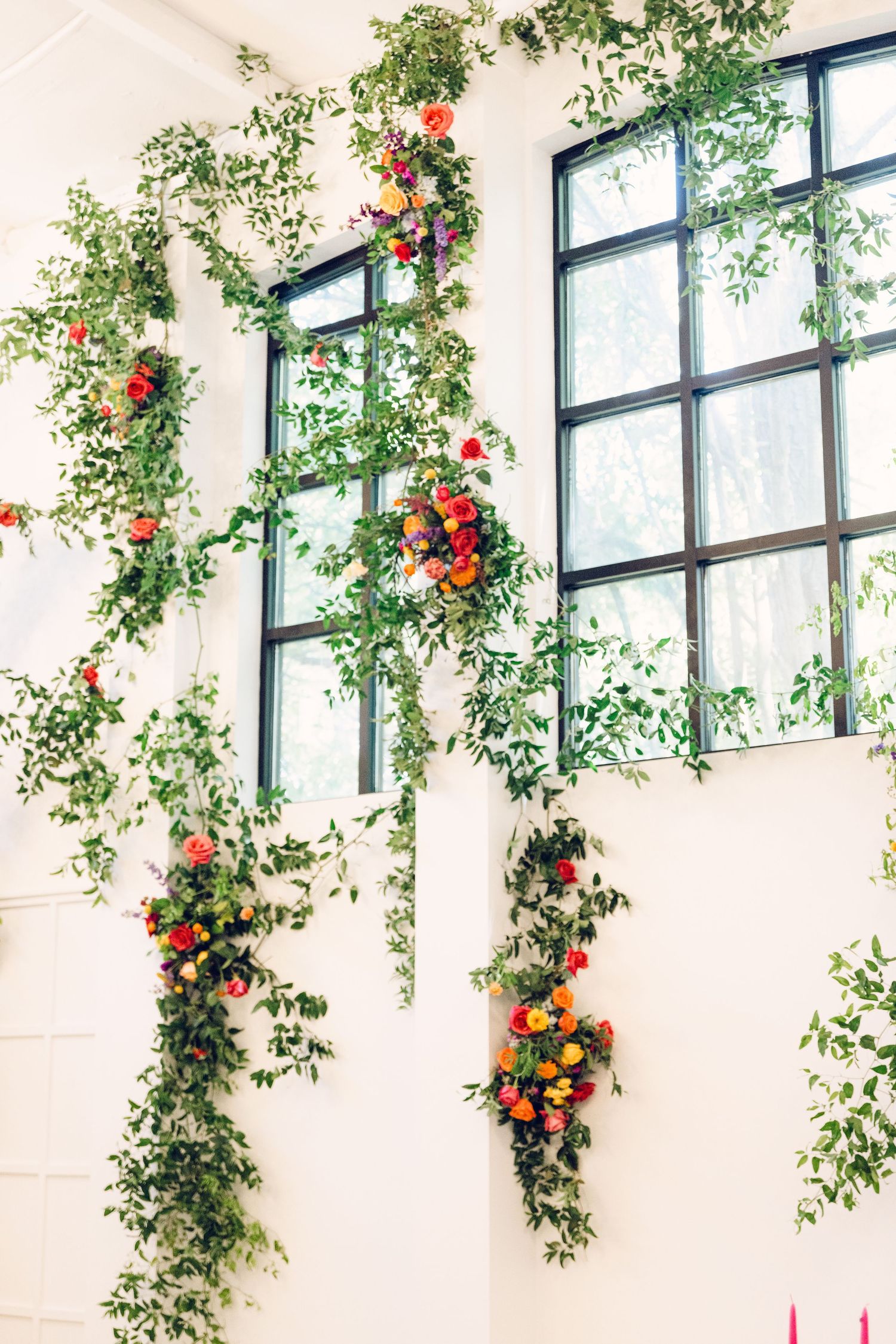 Elegant wedding venue interior with cascading greenery and red flowers hanging from windows in natural light.