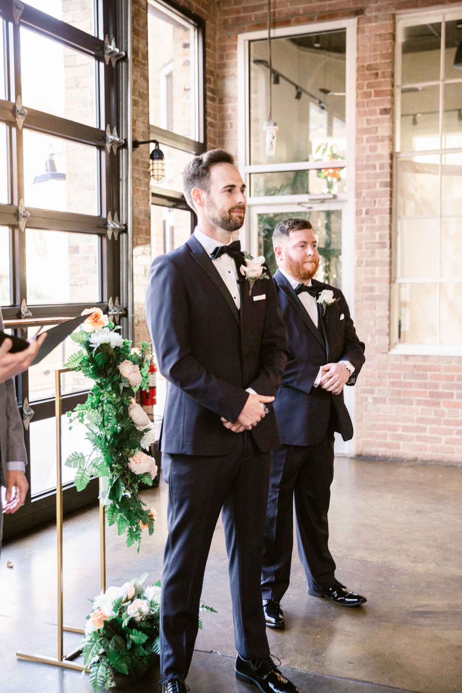 Groomsmen in black suits stand in a row inside an industrial brick venue decorated with green garlands.
